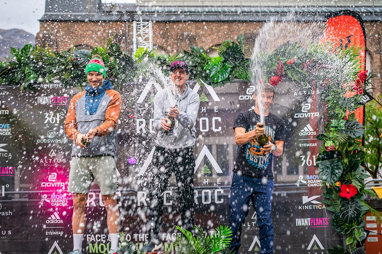 Three people stand on a podium, joyfully spraying champagne. The backdrop features logos and lush greenery. They wear casual clothing and appear to be celebrating a victory at an outdoor event.