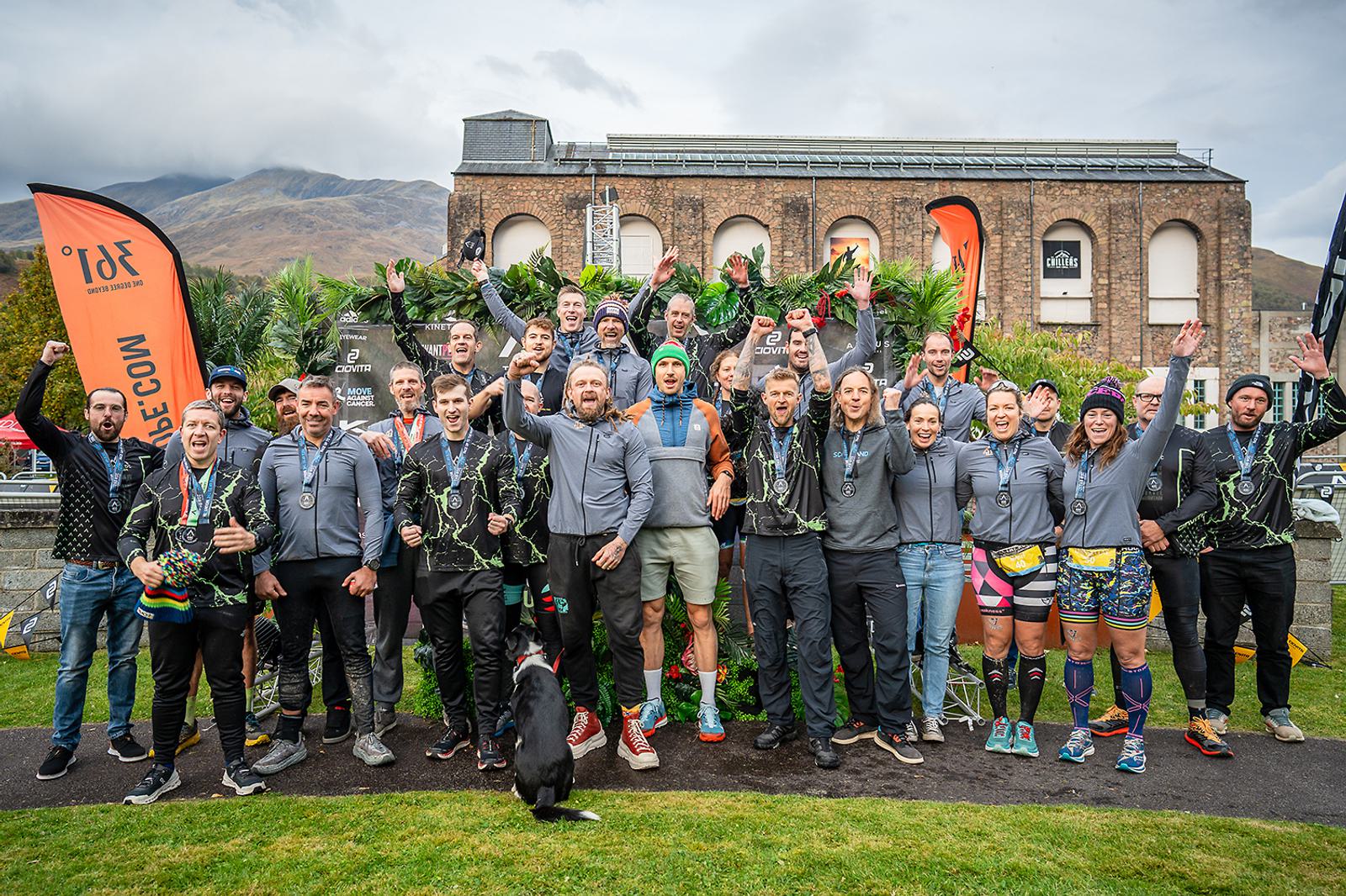 A group of people in athletic gear posing for a photo outdoors. They're standing on grass with a building in the background. Some hold medals and trophies, and a black dog is in front. Orange flags and greenery add to the scene. Everyone appears cheerful.