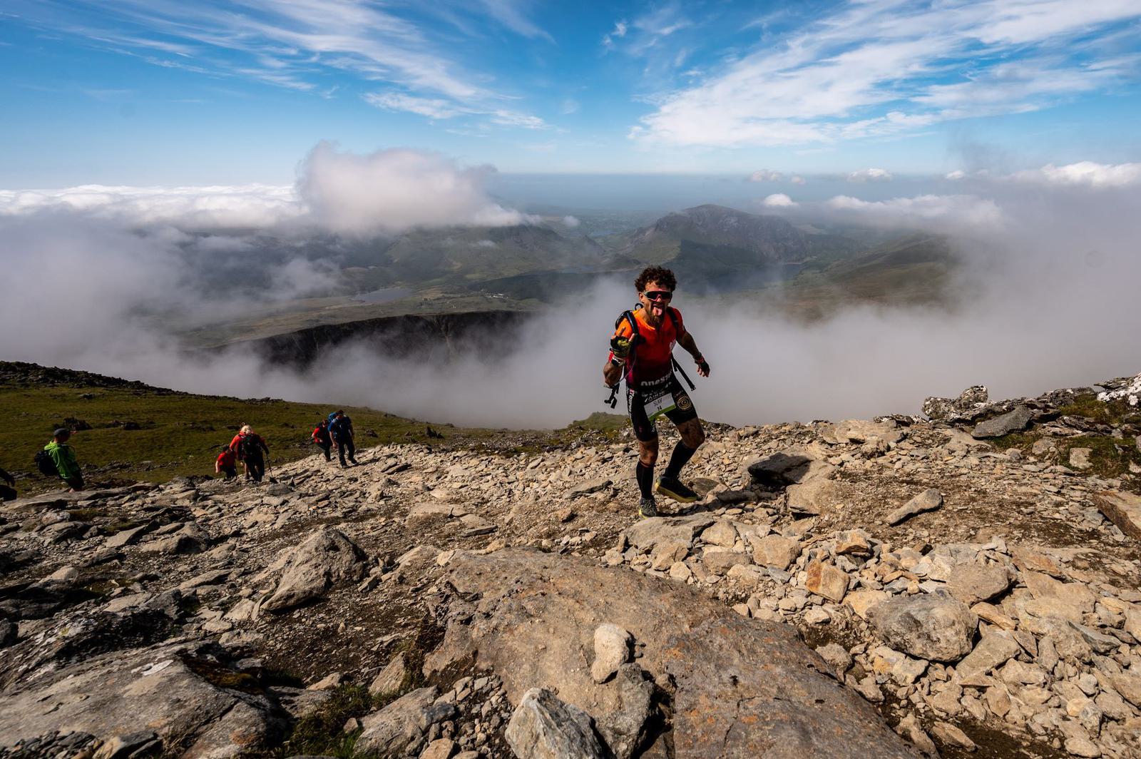 A group of hikers ascend a rocky mountain trail under a bright blue sky with scattered clouds. The leading person wears an orange jacket and green shorts, moving energetically ahead. Below them, vast landscapes of mountains and valleys stretch out, partially obscured by mist.