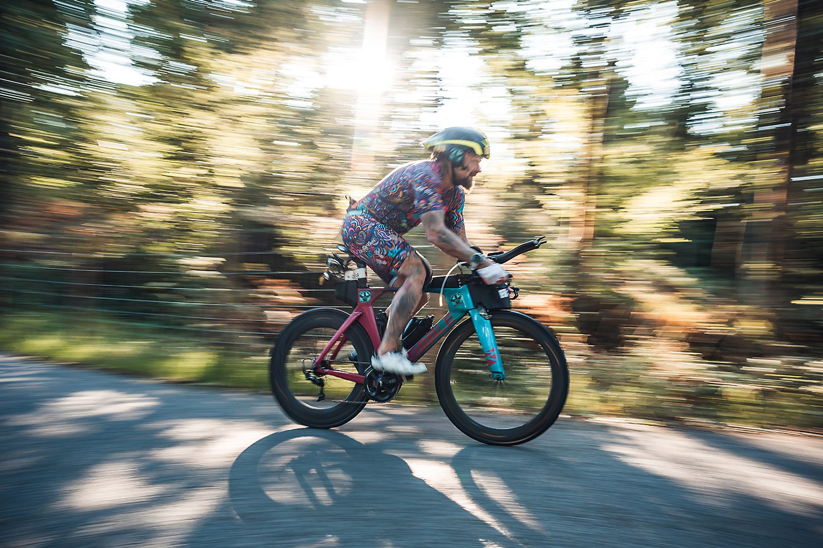 A cyclist in colorful attire and a helmet rides swiftly on a bicycle along a shaded forest path, with motion blur creating a sense of speed as sunlight filters through the trees in the background.