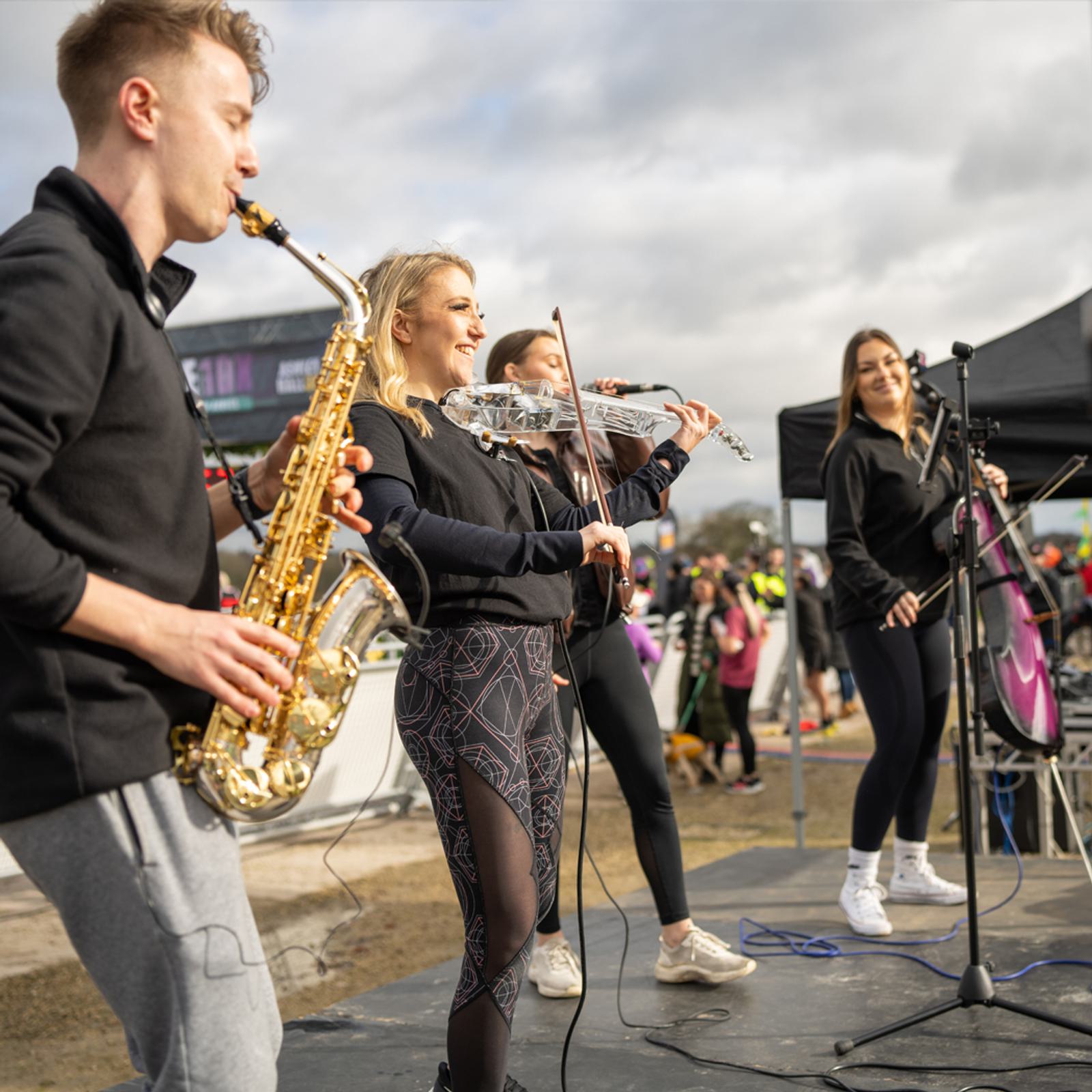 Four musicians perform on an outdoor stage. A man plays a saxophone, a woman plays a violin, another woman plays an electric violin, and a fourth woman sings into a microphone. The background shows a crowd of people and overcast skies.