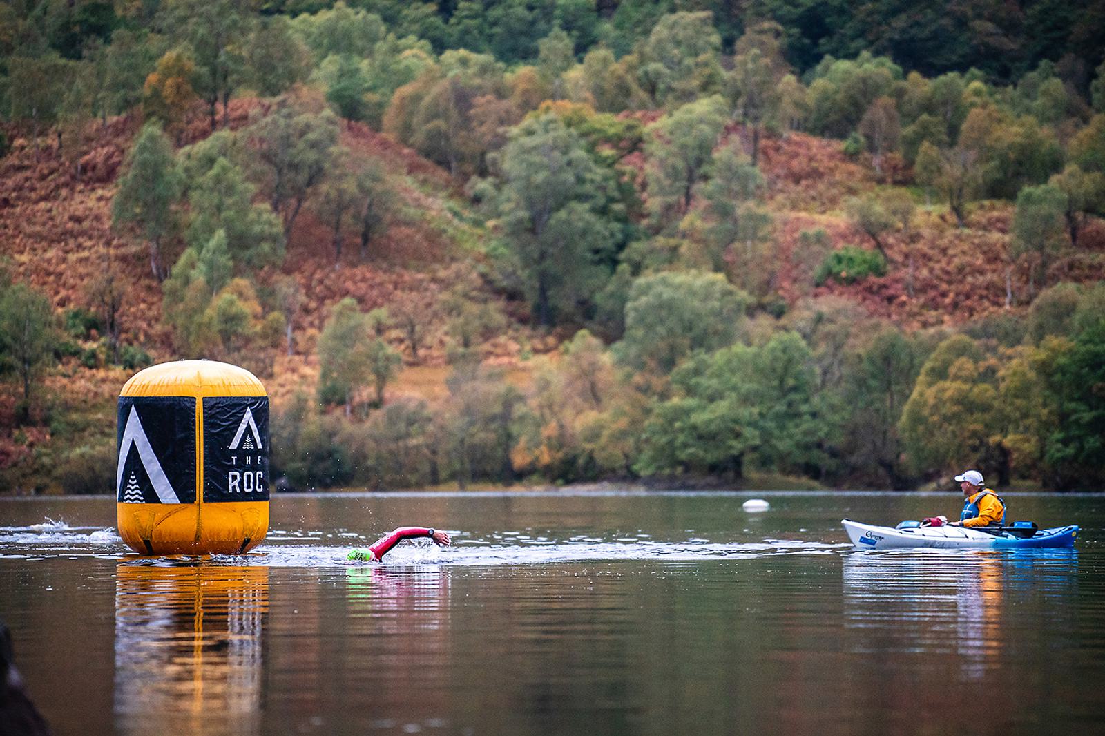 A person in a green and red wetsuit swims in a lake toward a large yellow buoy, while another person paddles a kayak nearby. The background consists of trees and rolling hills with autumn colors.