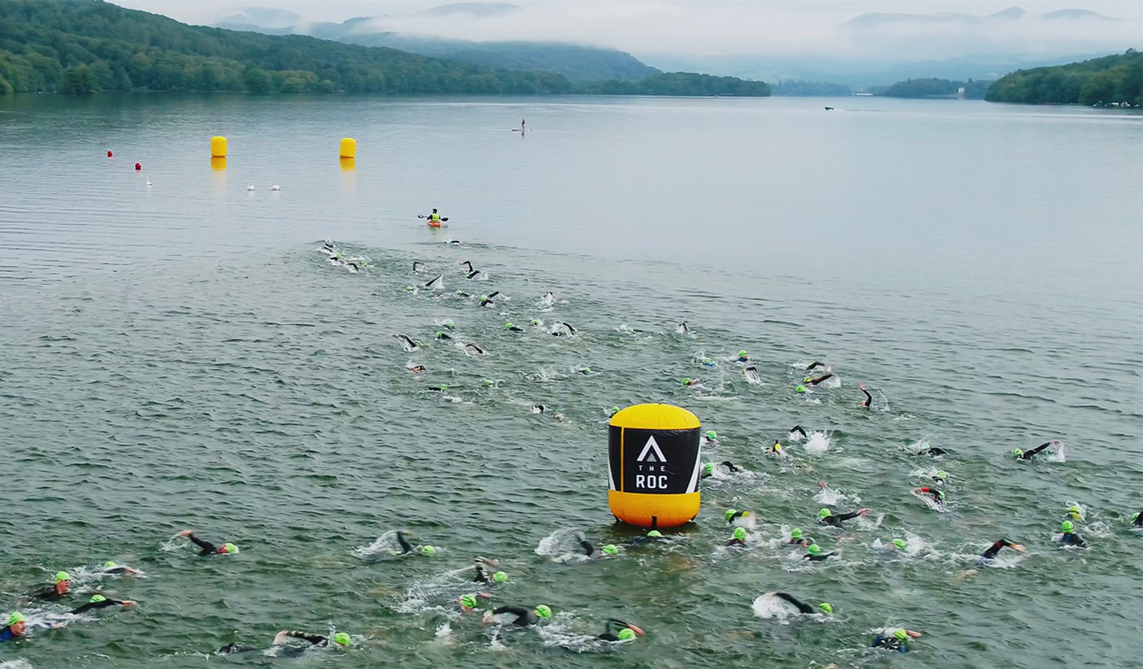 A large group of swimmers wearing green swim caps are participating in an open water race in a lake. Buoys with the logo "Roc" provide direction. The scene is set against a backdrop of misty mountains and dense forest under a cloudy sky.