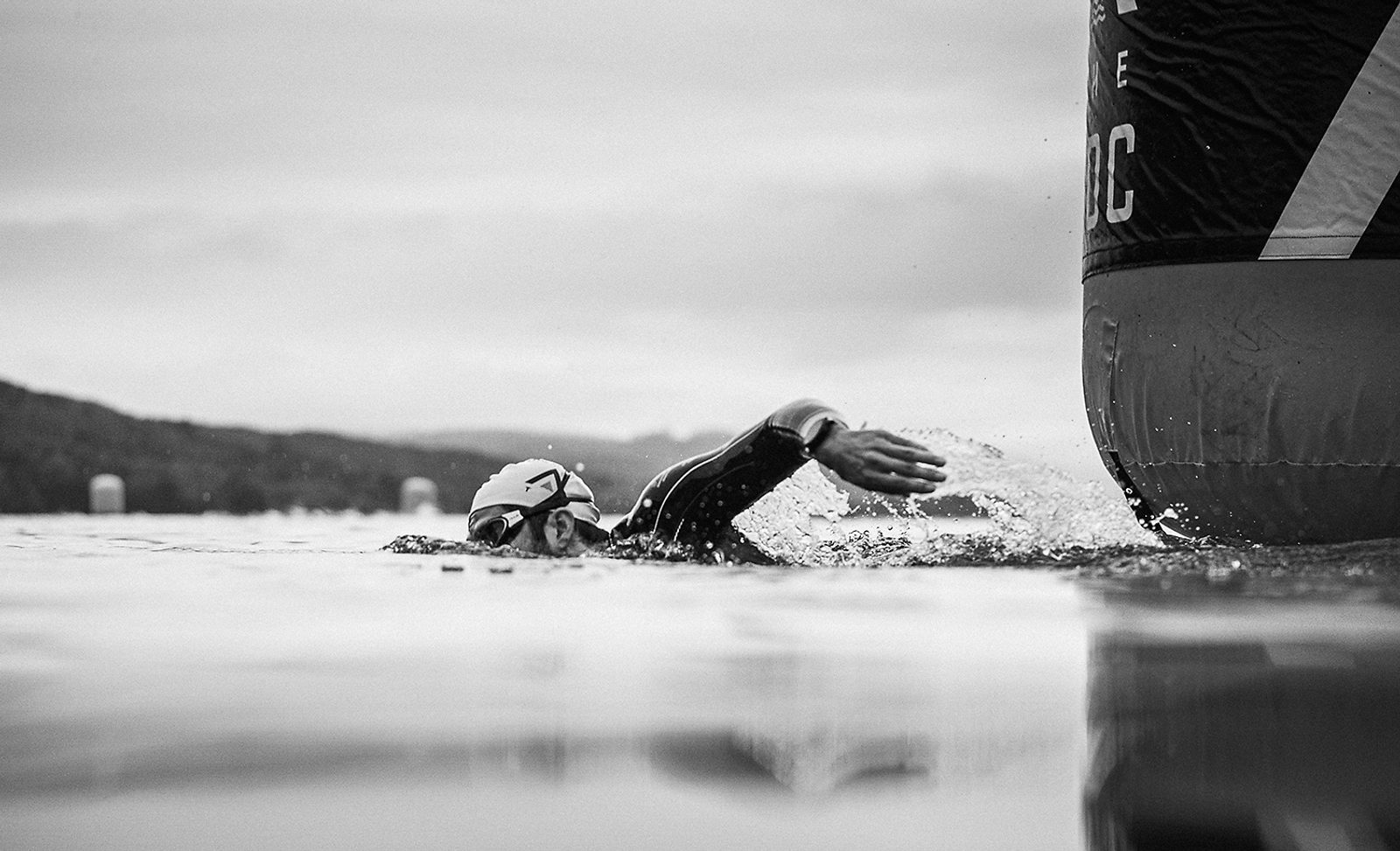 A black-and-white photo of a swimmer in a race turning around a large buoy in open water. The swimmer is wearing a swim cap and goggles, and the arm is outstretched mid-stroke. The background includes a hilly landscape and a cloudy sky.