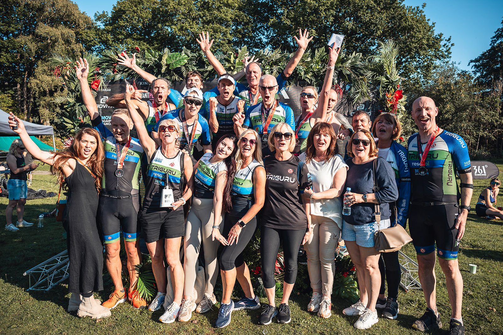 A group of cyclists and supporters celebrate outdoors, smiling and raising their hands in excitement. The cyclists are wearing matching gear with a green and blue design, and some have medals around their necks. Trees and event tents are visible in the background.