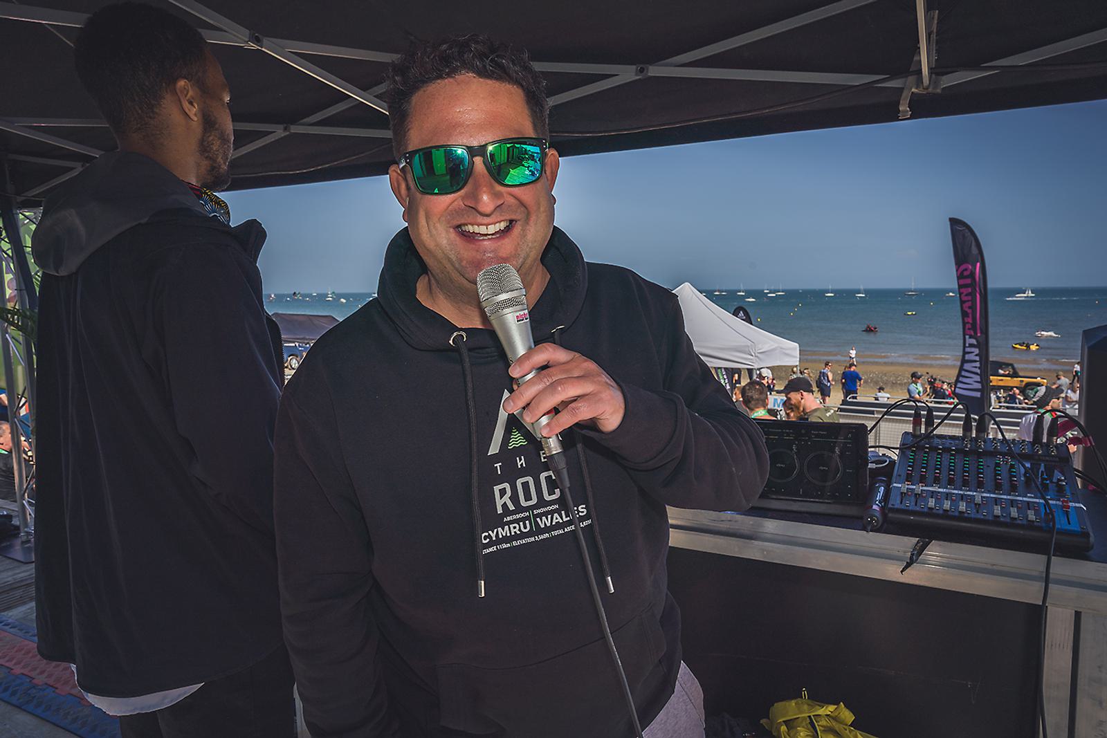 A man wearing sunglasses and a black hoodie holds a microphone and smiles under a canopy at an outdoor beach event. Another person stands behind him, and sound equipment is visible on a table. The scene includes a blue sky and people enjoying the beach in the background.