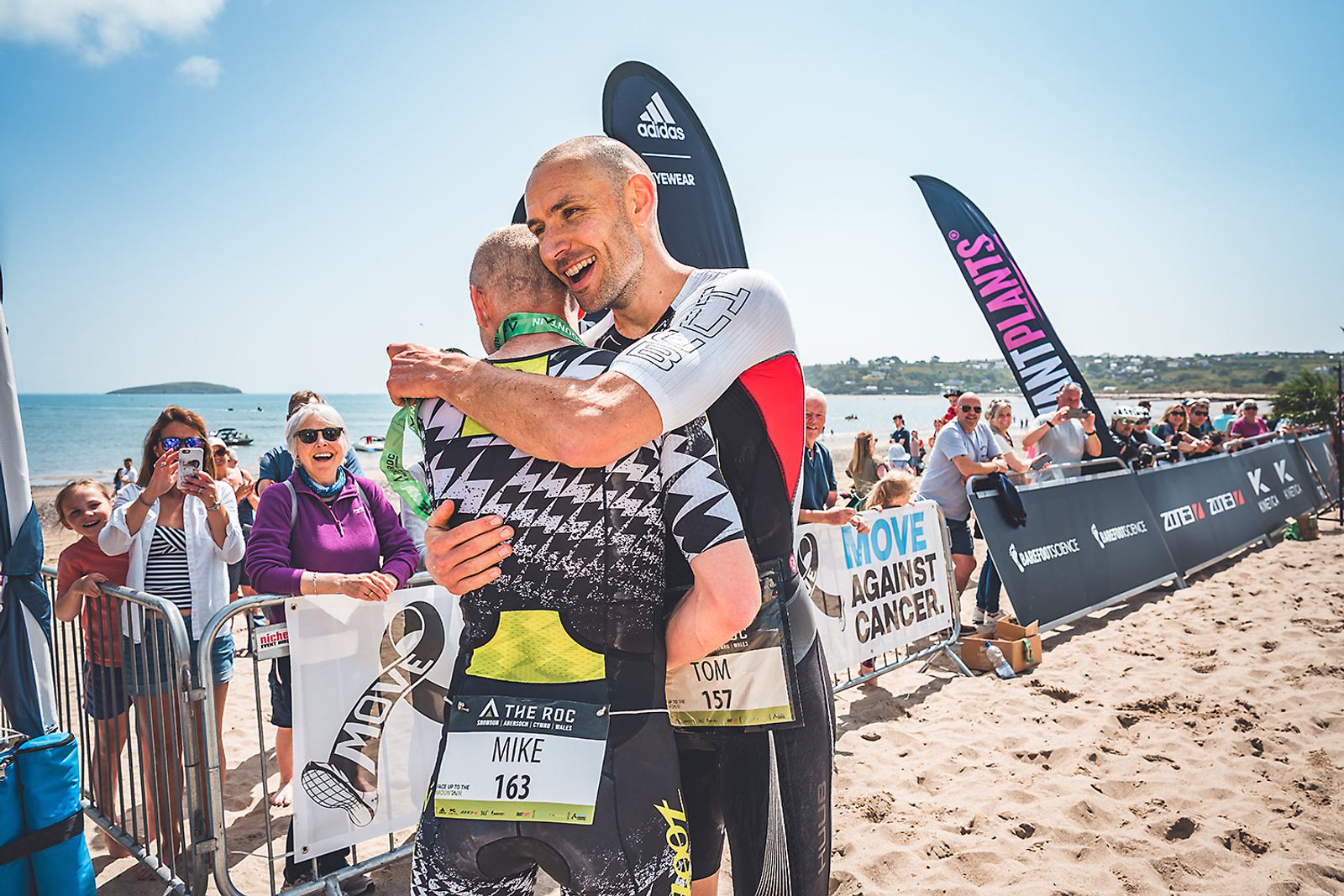 Two triathletes embrace at the finish line on a sandy beach. One athlete, wearing a black and green suit with "Mike 163" on the back, is being hugged by another athlete in a white and black suit, "Tom 157." Spectators and event banners are visible in the background.