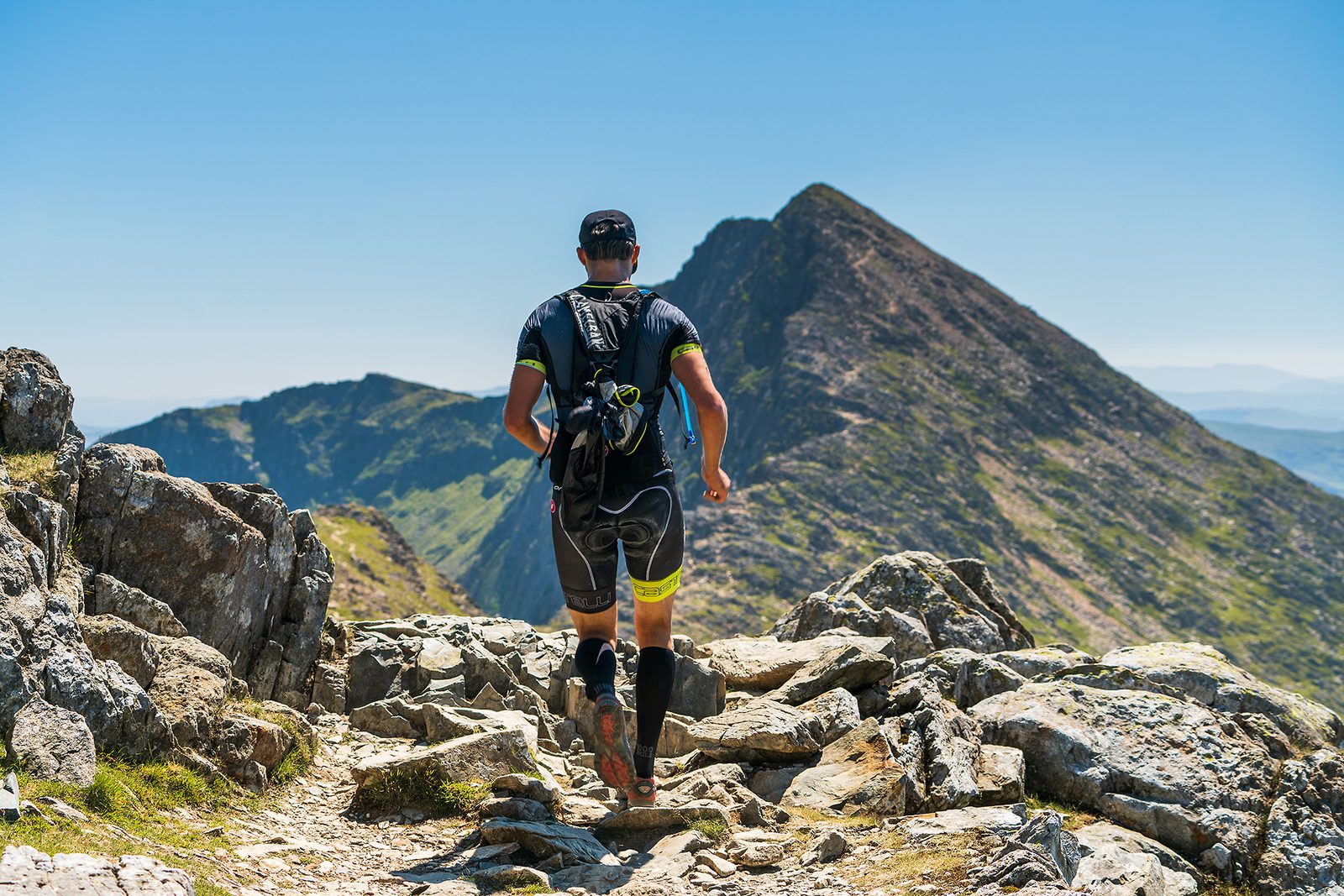 A person wearing a backpack and athletic gear is running on a rocky mountain trail. Ahead, a mountain peak towers against a clear blue sky. The trail descends slightly before ascending towards the peak. The surrounding landscape is rugged and mountainous.