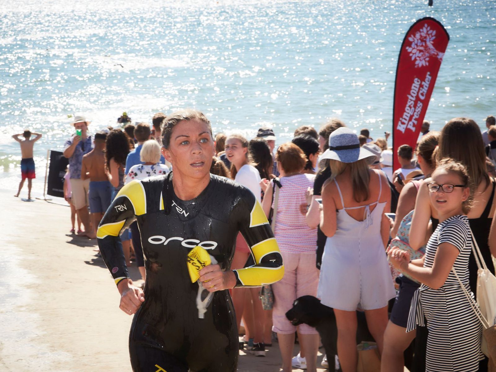 A person in a wetsuit runs up a ramp from the water during a triathlon event. Crowds of spectators line both sides of the ramp, watching and cheering. The sun shines brightly, reflecting on the ocean in the background. A red flag labeled "Lighthouse Triathlon" is visible.