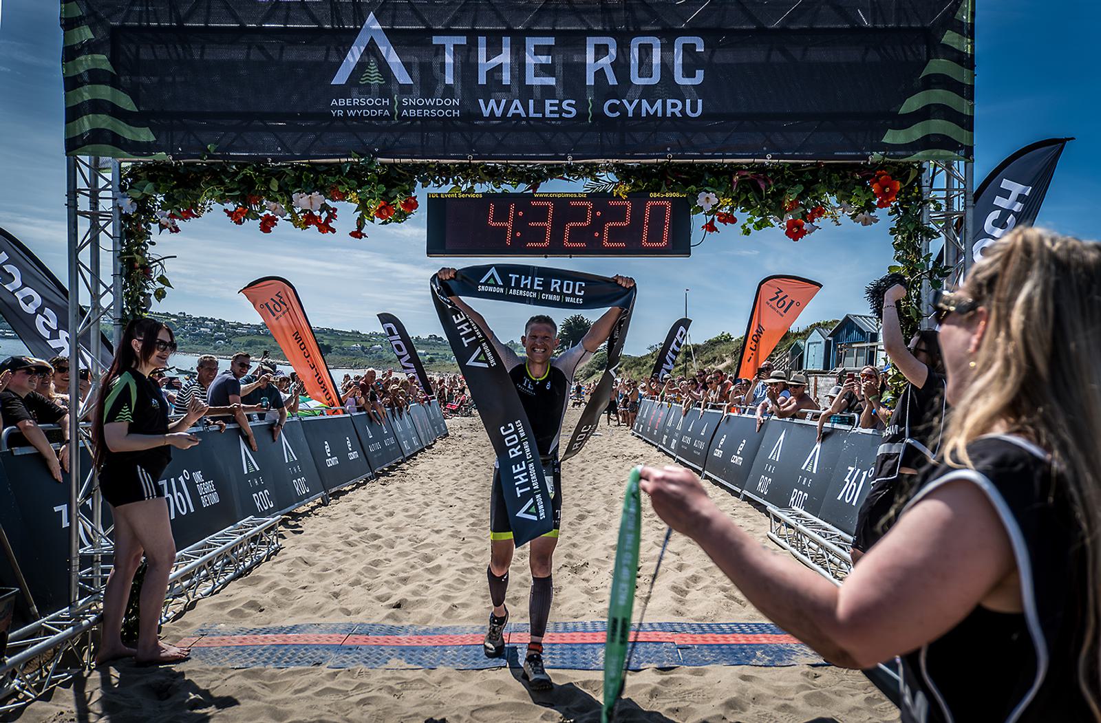 A triathlete crosses the finish line at The ROC Wales, holding a banner triumphantly. The time displayed above is 4:32:20. Spectators cheer on both sides under sunny skies.