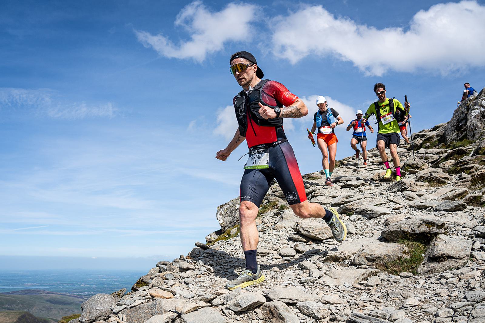 Athletes run down a rocky mountain trail in a race. The leading runner wears a red and black outfit with sunglasses. The sky is clear and blue, and distant landscapes are visible in the background.