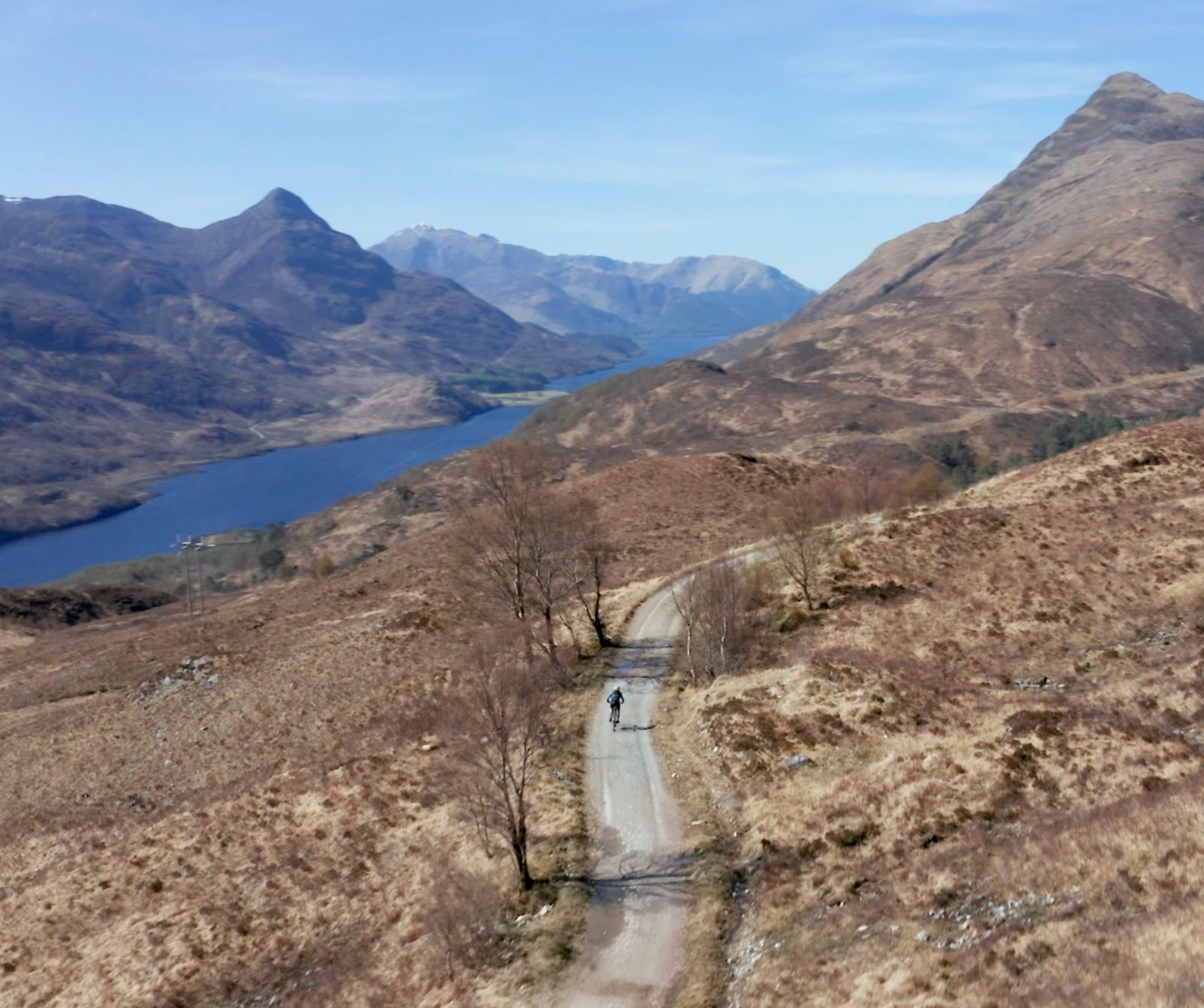 A lone cyclist rides on a winding dirt road flanked by bare, brown hills, with a distant blue lake and rugged mountains under a clear blue sky in the background. Sparse trees are scattered along the landscape.
