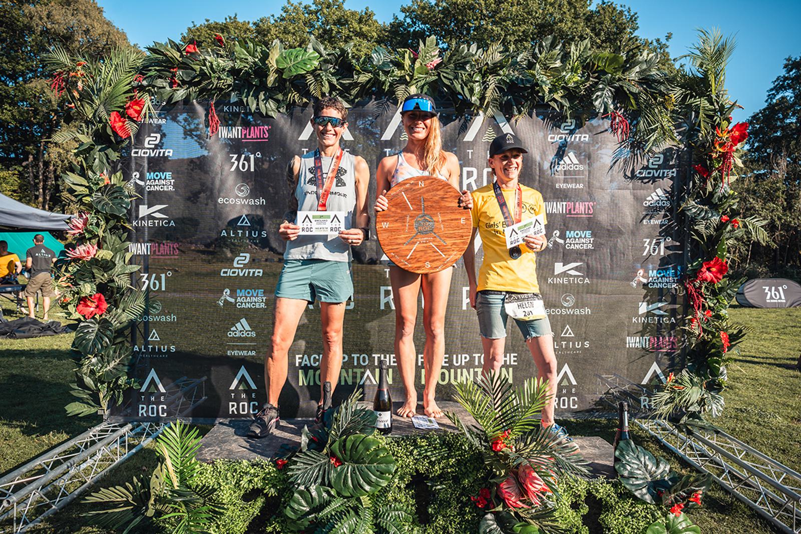 Three athletes stand on a podium adorned with lush greenery and flowers, holding their awards after a race. The backdrop features sponsor logos and the event name. The central figure, a woman, lifts a trophy above her head. The sunny weather highlights their accomplishment.