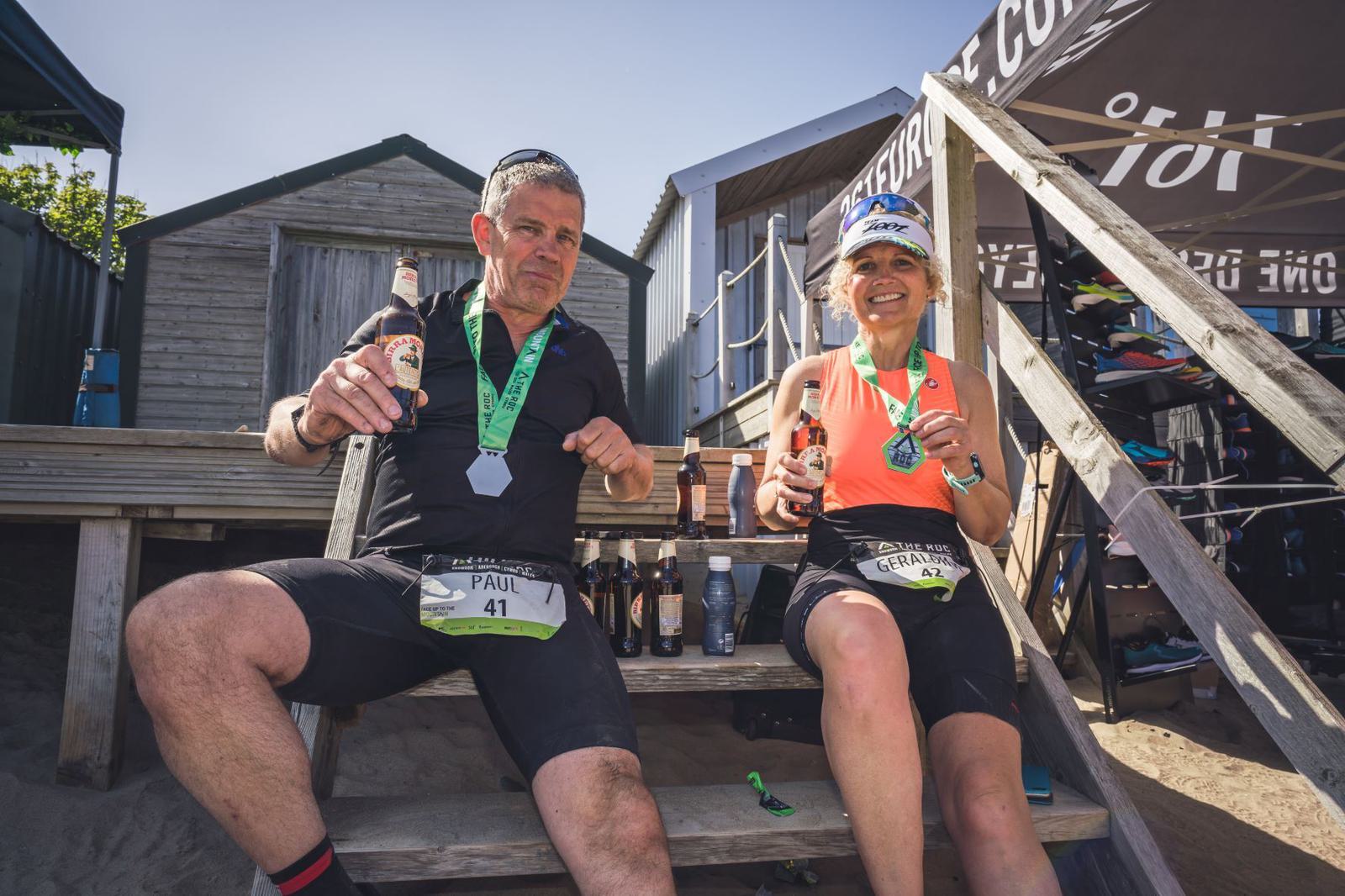 Two cyclists, a man and a woman, are sitting on wooden steps, each holding a beer and wearing medals around their necks. They both have race bibs, the man's reads "PAUL" and the woman's reads "GRACE". They appear to be celebrating post-race.