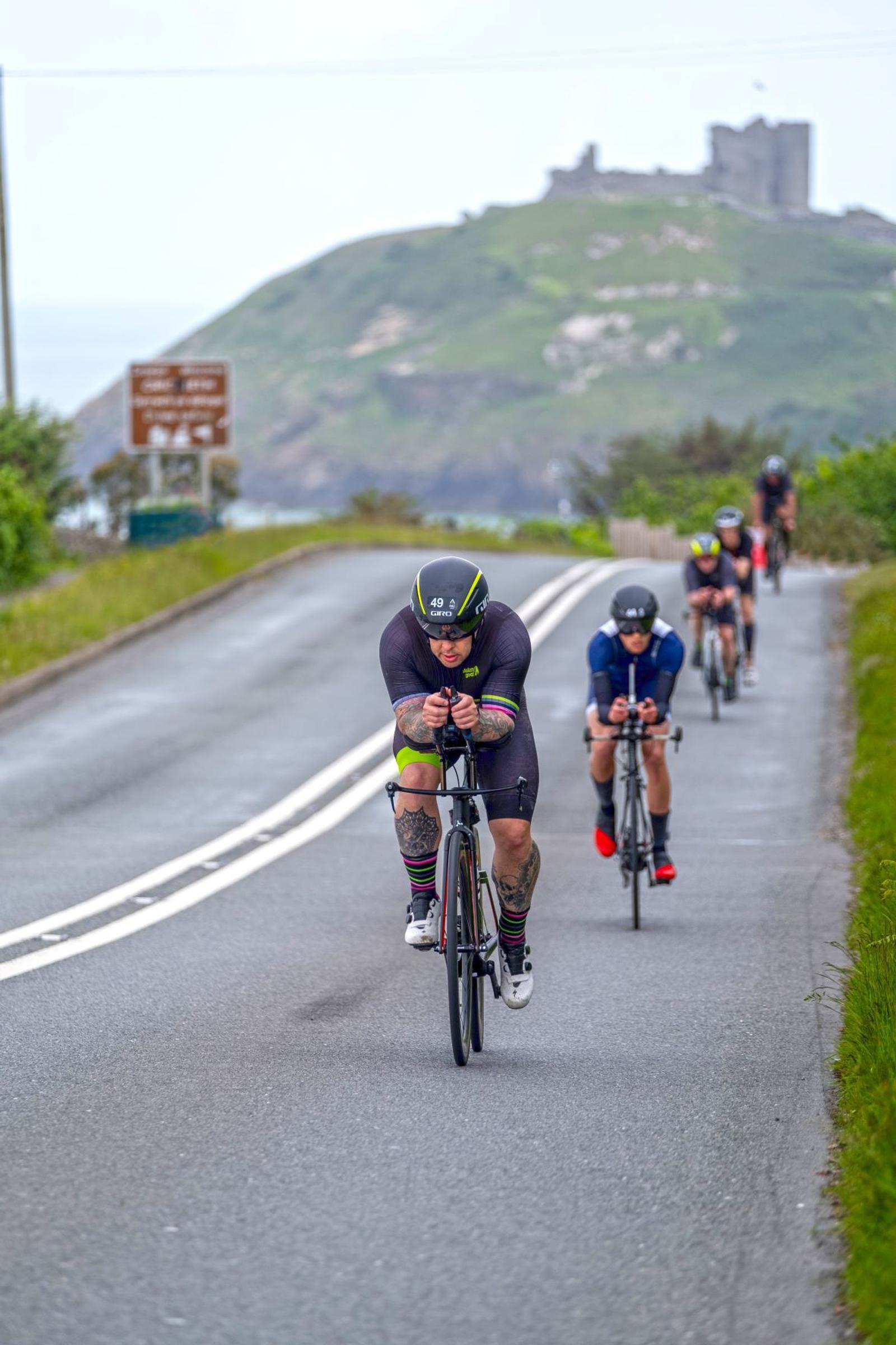 A group of cyclists riding on a two-lane road with a grassy hill and a castle in the background. The leading cyclist is wearing a black helmet and cycling gear. The road is flanked by greenery and the sky is overcast.