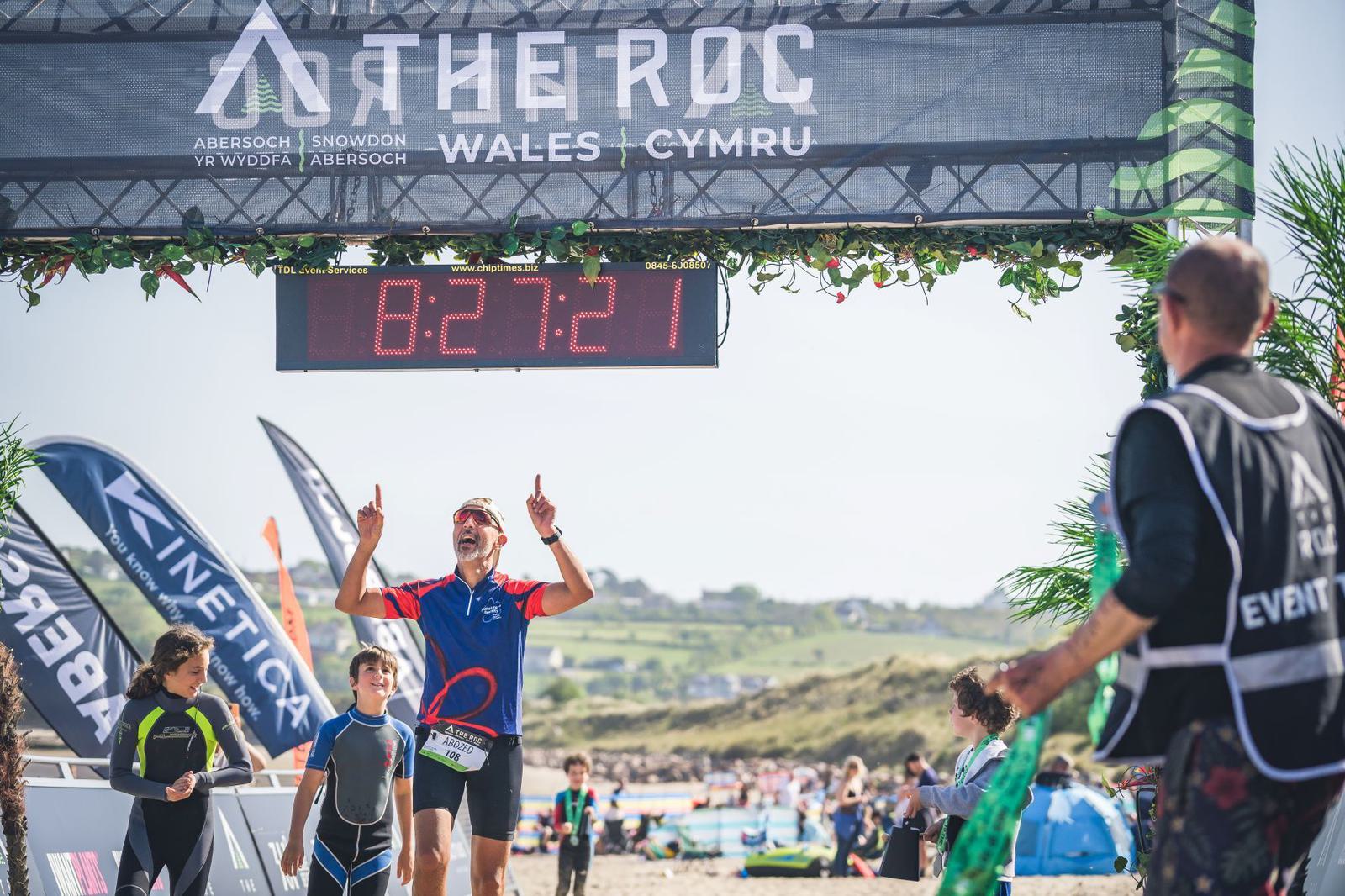A male athlete crosses the finish line of a race at "The ROC Wales" triathlon, visibly elated with arms raised and fingers pointing upwards. The timer above displays 8:27:21. Spectators, other participants, and event staff are seen in the background.