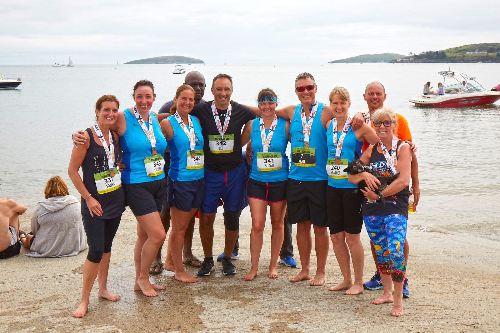 A group of ten people in running attire stands on a beach, smiling and wearing medals. Some have their arms around each other. The ocean and boats are visible in the background. One person holds a small dog.