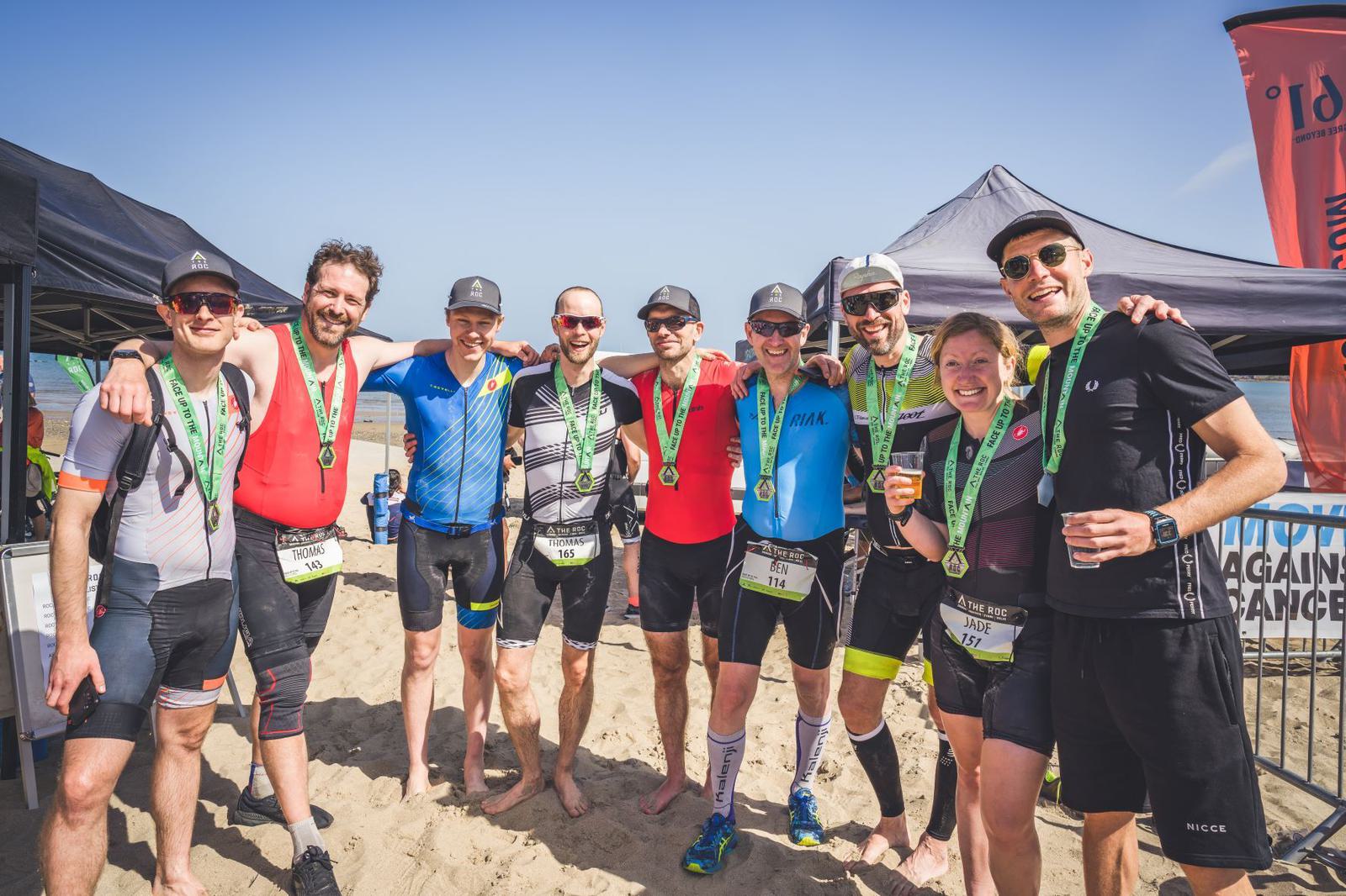 A group of eight people stand on a sandy beach wearing cycling and running gear, each displaying medals around their necks. They appear cheerful, some holding drinks. Behind them are tents and signage from the event, with a bright blue sky overhead.