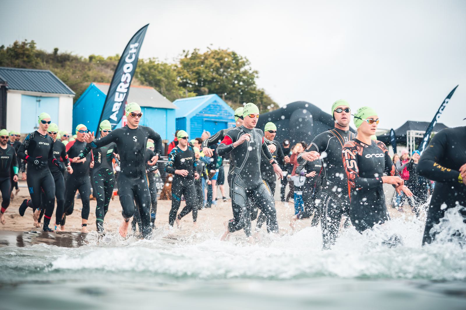 A group of triathletes in black wetsuits and green swim caps run into the ocean from a sandy beach, participating in a triathlon event. Spectators and beach huts are visible in the background under a cloudy sky.