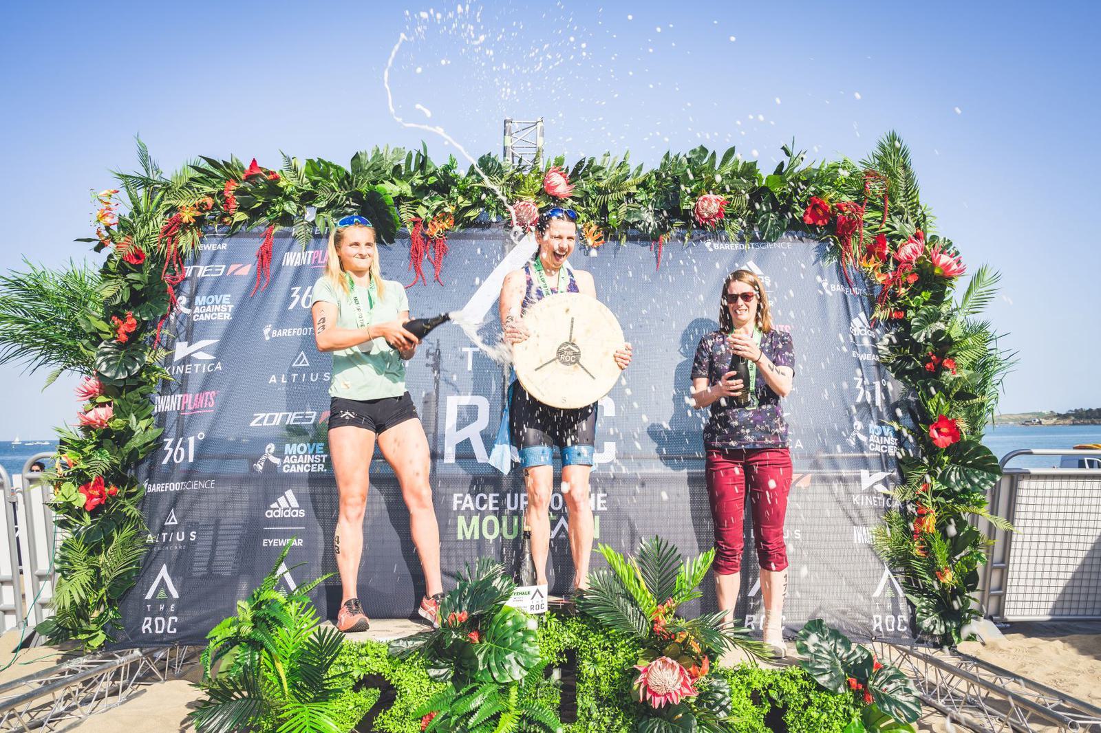 Three athletes stand on a podium decorated with a floral arch and a backdrop displaying various logos. The athlete in the center, holding a trophy, is being sprayed with liquid. The two athletes on either side hold their awards and smile. A beach can be seen in the background.