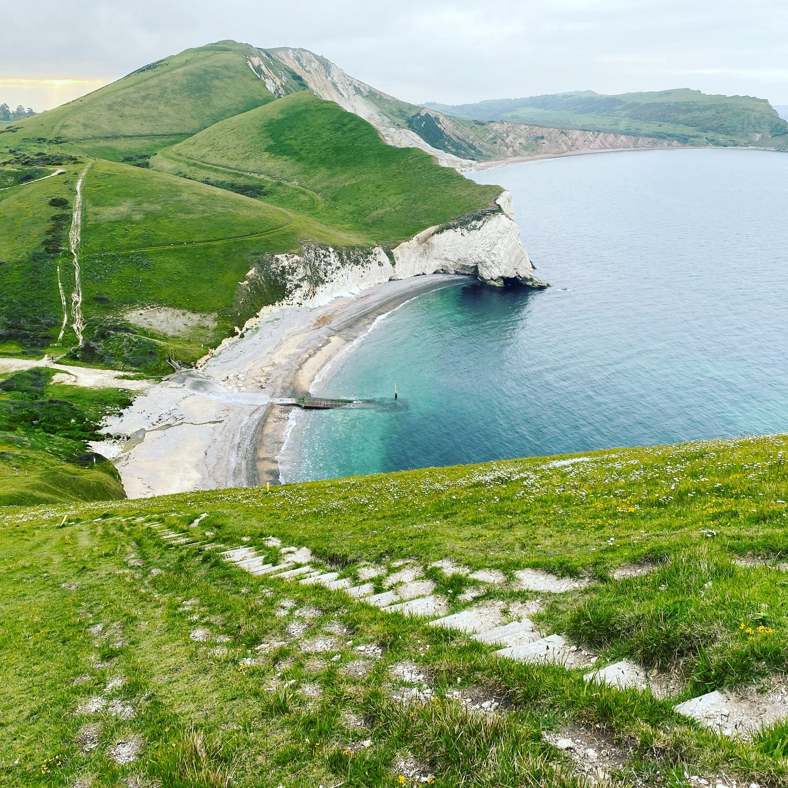 A narrow path leads down a grassy hillside to a scenic horseshoe-shaped bay with calm, turquoise water. White cliffs and green hills surround the bay. The sky is cloudy, and wildflowers dot the hillside.