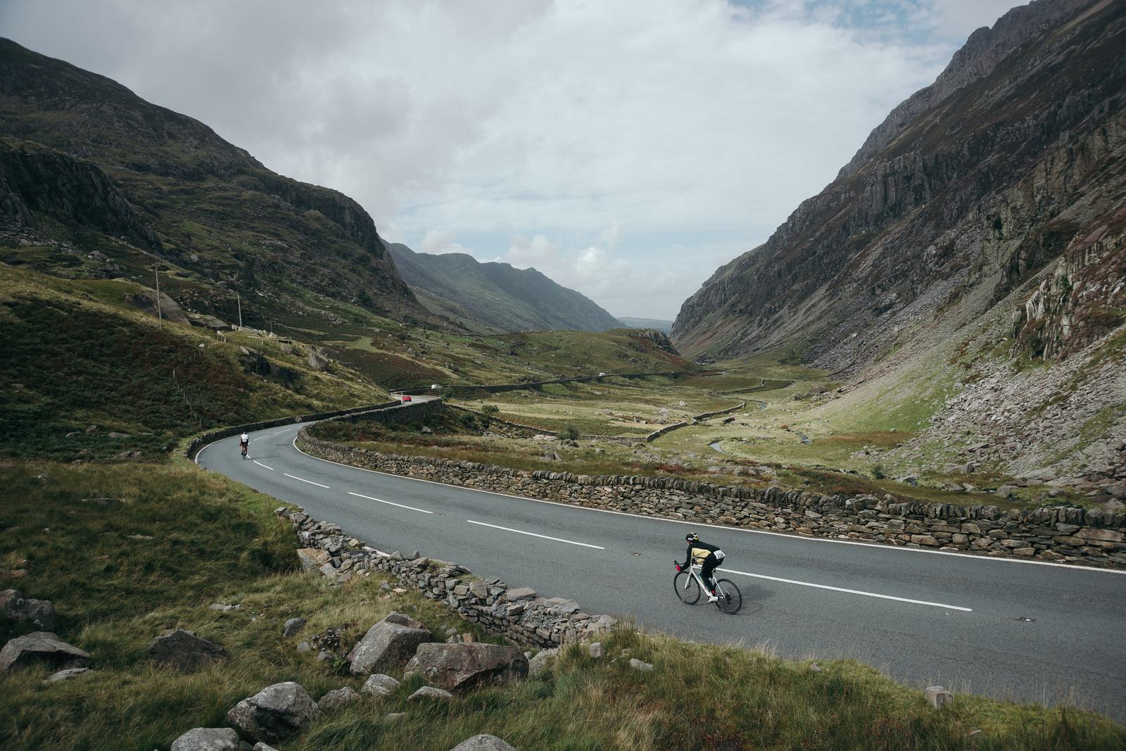 A person rides a bicycle on a winding road surrounded by tall, rocky hills and a lush, green valley. Another cyclist can be seen in the background. The sky is partly cloudy, casting a soft light on the serene landscape.