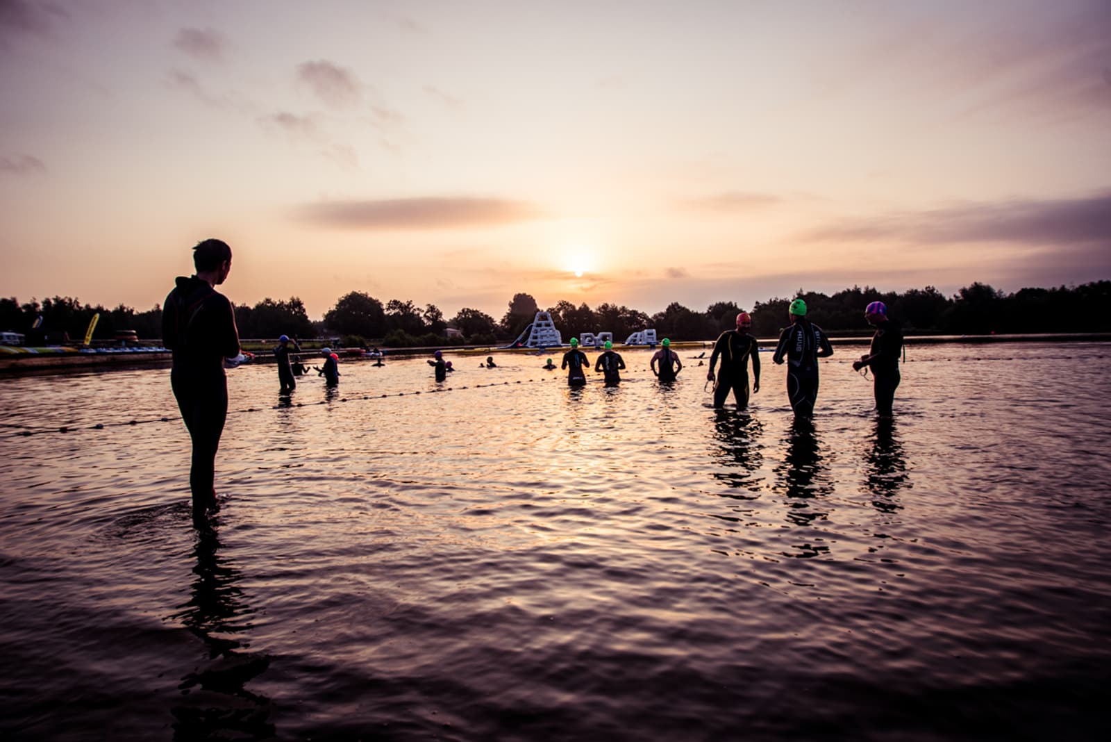 A group of people in wetsuits and colorful swim caps stand in shallow water at sunrise, preparing for a swim. A lifeguard or coach stands to the left, overseeing the participants, with trees and an inflatable structure visible in the background.