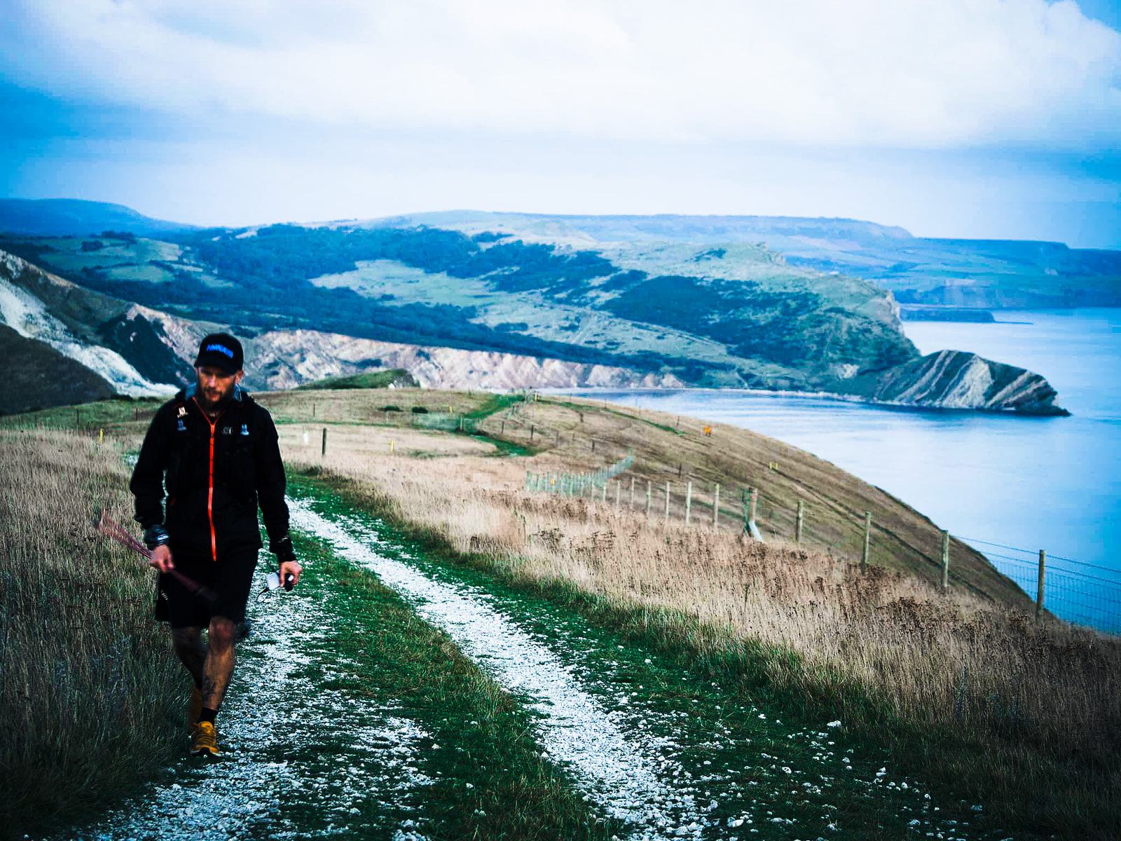 A person wearing a cap, jacket, shorts, and running shoes walks along a dirt path on a cliffside. The path overlooks the sea and rolling green hills under a cloudy sky. The background features dramatic coastal scenery with fenced-off sections and rugged terrain.