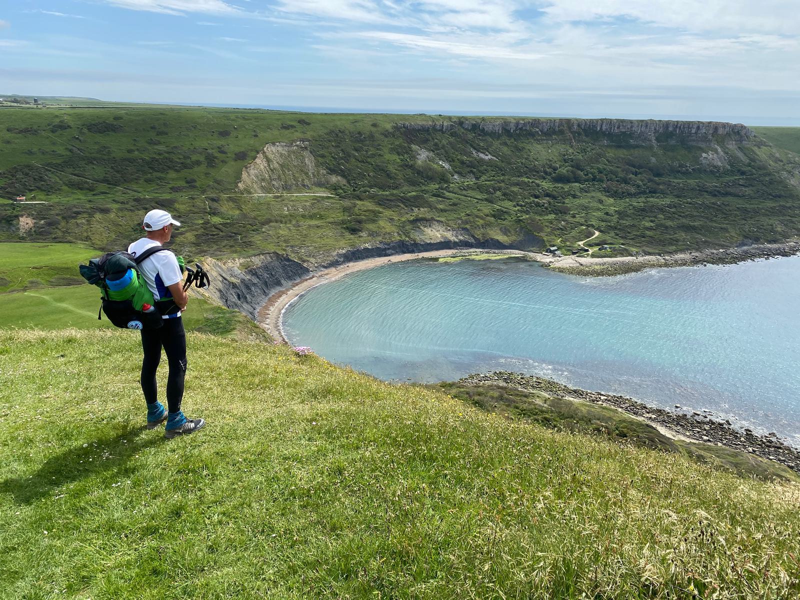A person wearing hiking gear stands on a grassy hilltop, holding walking sticks and looking out at a scenic coastal view with cliffs, green terrain, and a calm blue ocean. The sky is partly cloudy, casting soft light on the landscape.