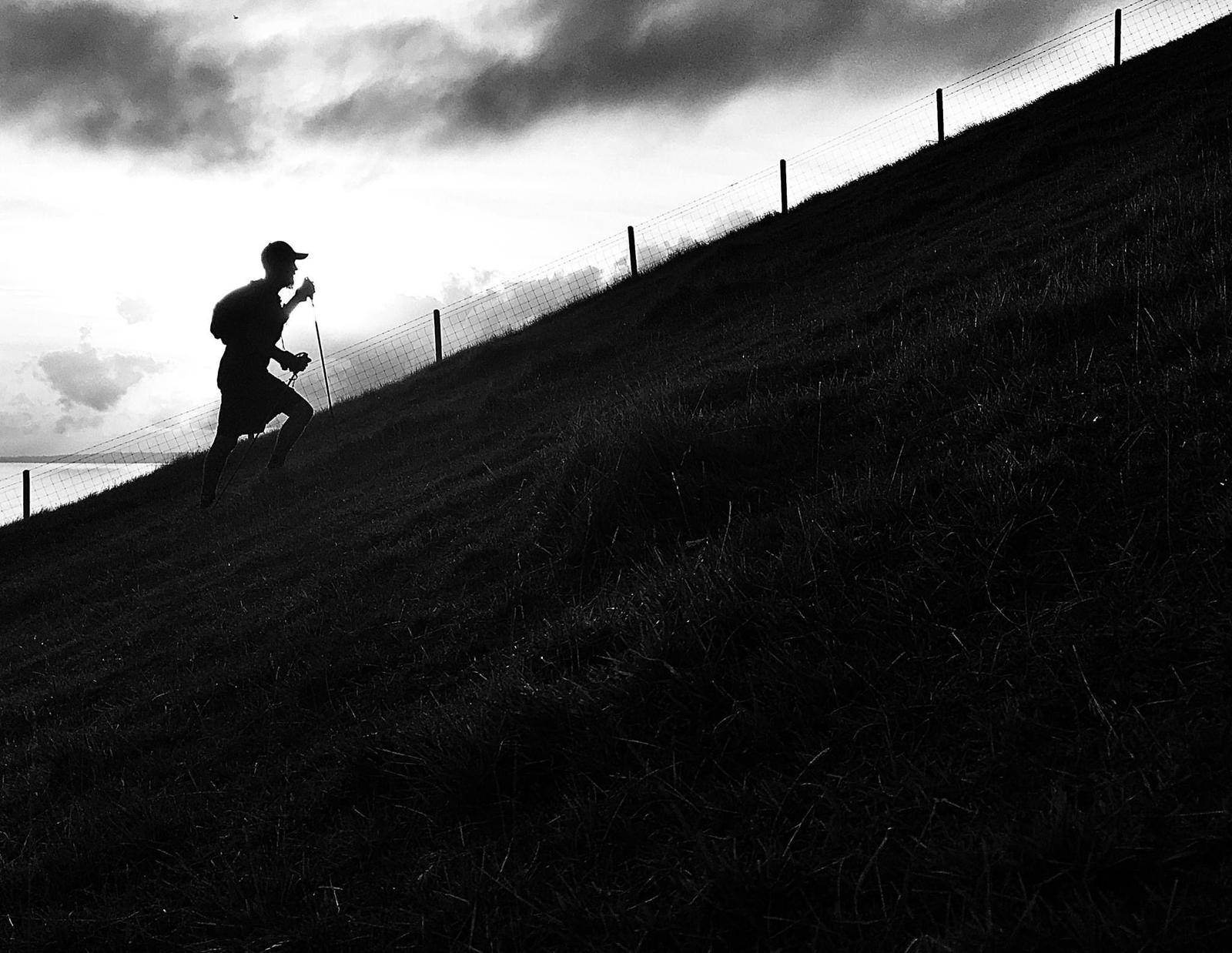 A silhouette of a person running uphill on a grassy slope against a cloudy sky. They are wearing a cap and carrying a backpack, with a fence running parallel to their path. The image is in black and white.