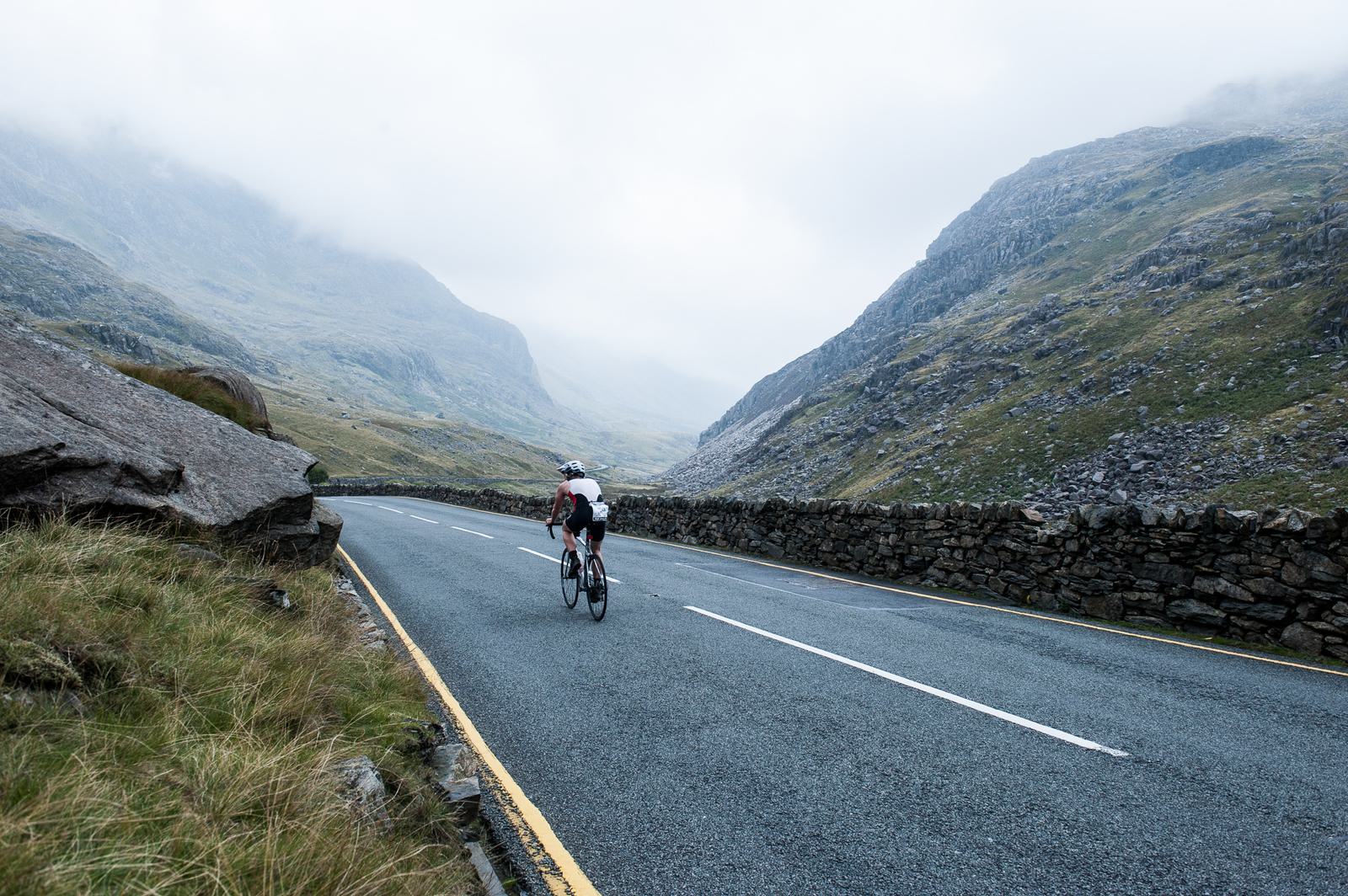 A cyclist rides along a winding mountain road flanked by stone walls and mist-covered hills. The scene is overcast, with gray skies and a sense of isolation in the expansive, rugged landscape.