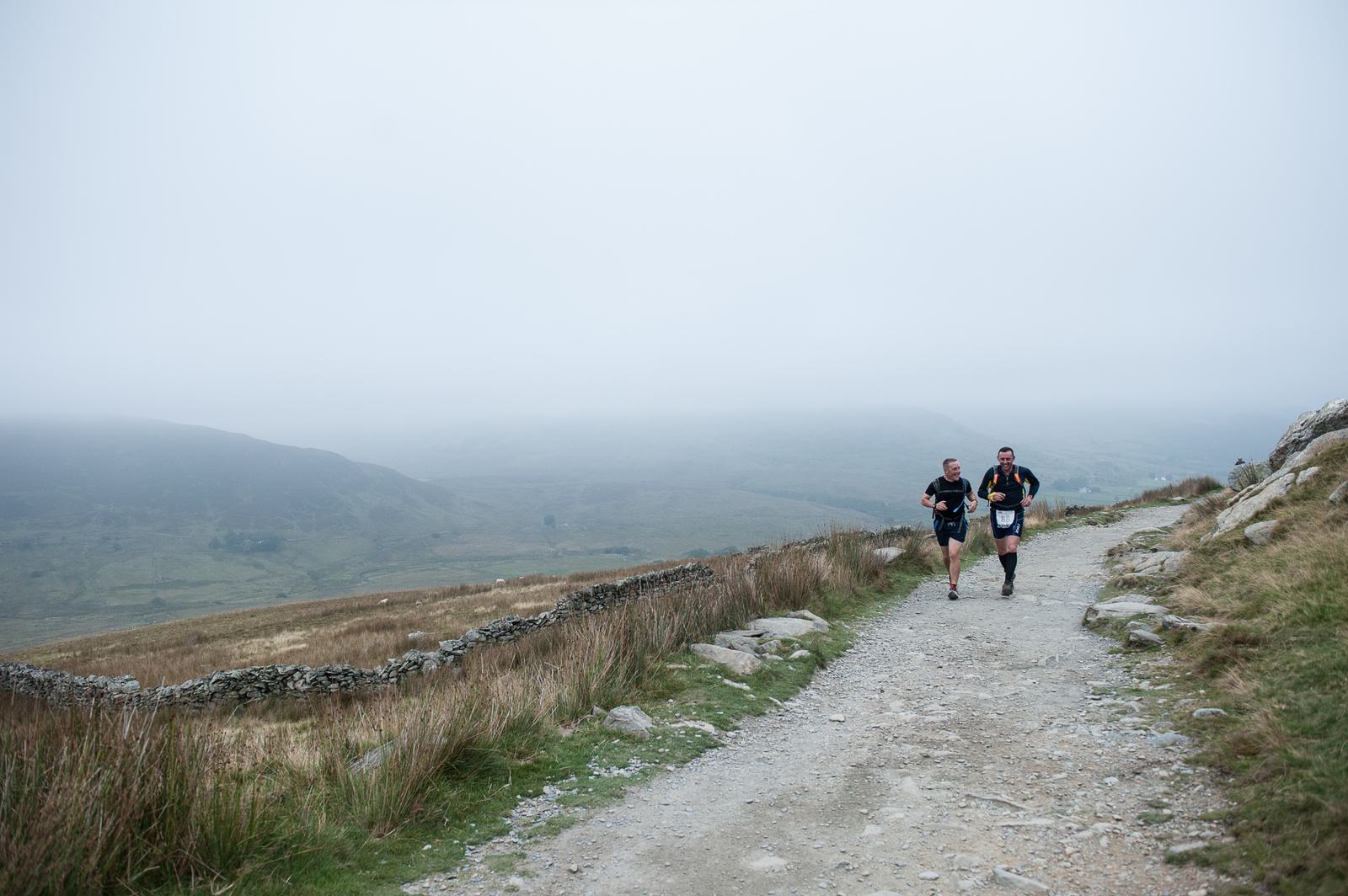 Two runners in athletic gear jog along a rocky trail surrounded by grassy terrain and a stone wall on a foggy day. The landscape in the background is misty and expansive, suggesting they are in a remote or rural area.