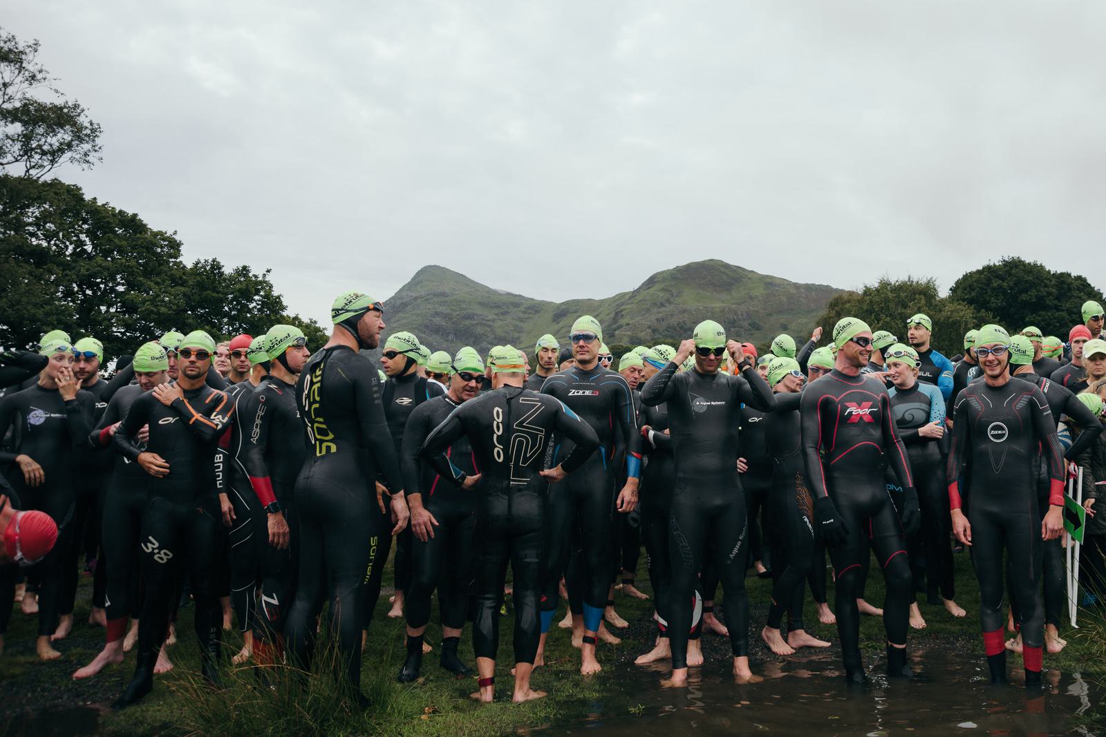 A large group of swimmers in wetsuits and green swim caps stand together outdoors, preparing for an open-water swim event. Mountains and cloudy sky form the backdrop, with some participants at the front of the group adjusting their gear and interacting with each other.