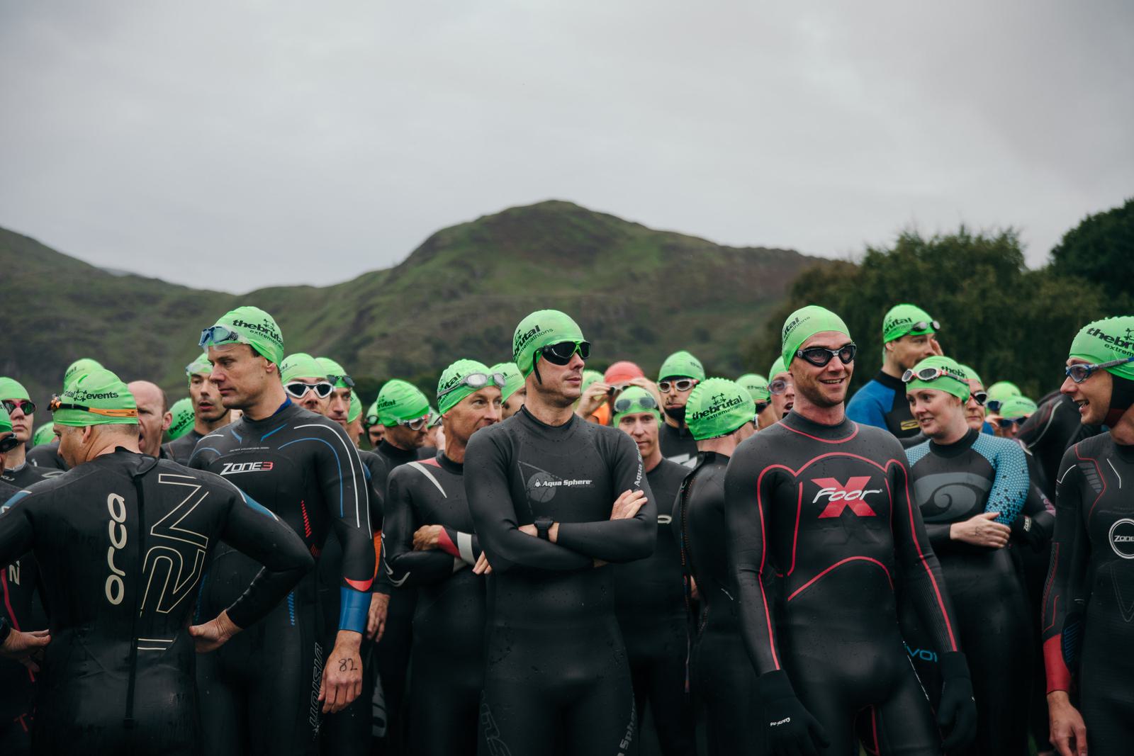 A group of swimmers wearing wetsuits and bright green swim caps gather at the starting line of a race. They stand in front of a mountainous landscape under an overcast sky, some looking serious and focused while others chat with one another.