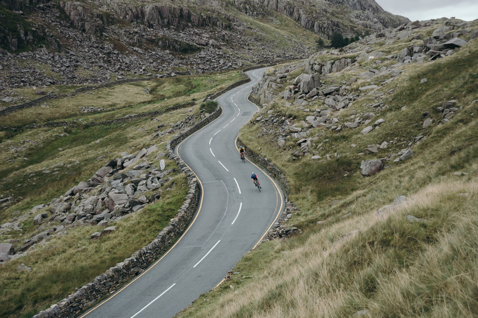 A winding road snakes through a mountainous landscape. Two cyclists are pedaling uphill, surrounded by grassy slopes and rocky terrain. The sky above is overcast, adding a dramatic feel to the rugged scene.