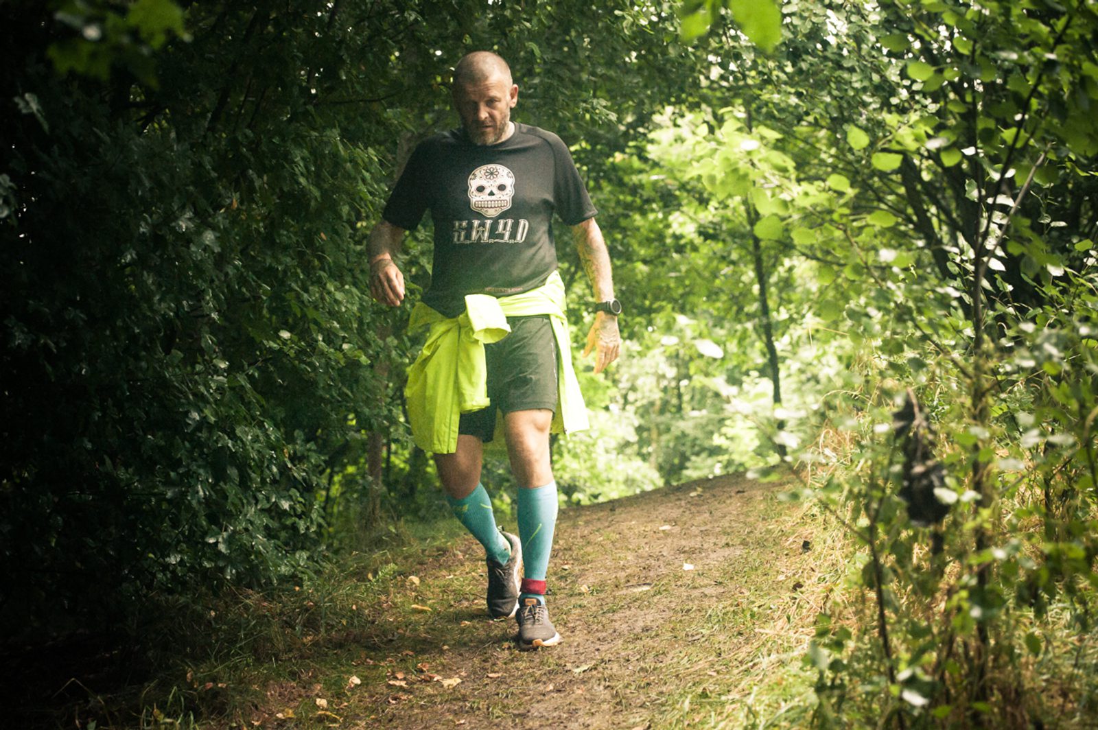 A person is walking on a dirt path through a lush, green forest. They are wearing a black T-shirt with a graphic design, black shorts, and bright blue knee-high socks. A neon yellow jacket is tied around their waist. The path is surrounded by dense foliage.