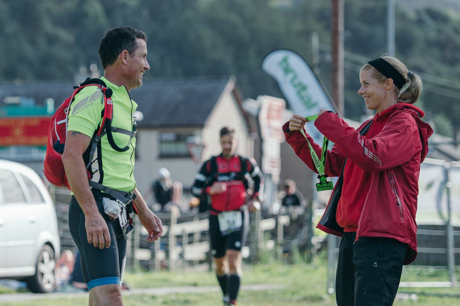 A man in athletic gear with a red backpack stands smiling at a woman in a red jacket who is handing him a medal. Both are outdoors, and there is a runner in the background along with some buildings and a banner visible.
