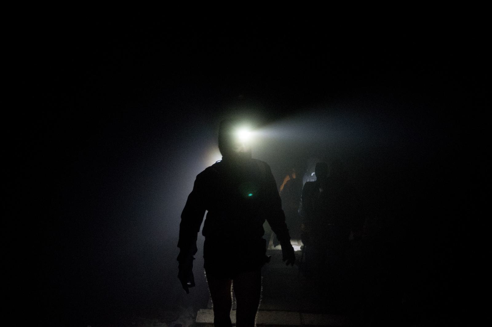 A person wearing a headlamp walks in the darkness, casting a bright beam of light. Silhouettes of other individuals can be seen following behind, shrouded in low light and shadows. The atmosphere is mysterious and dimly lit.