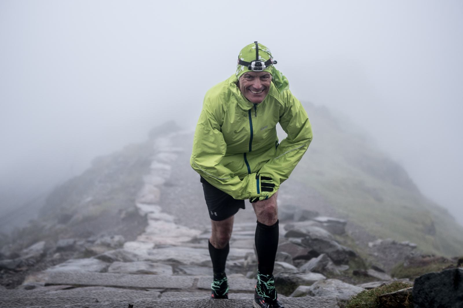 A person wearing a bright green jacket, headlamp, black shorts, and knee-high socks is hiking up a rocky trail on a foggy day. The individual is bending slightly forward and smiling, with mist and rocks visible in the background.