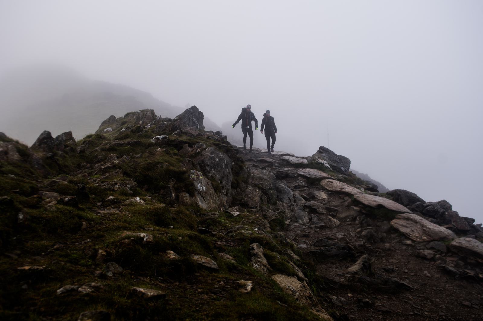 Two hikers walk along a rocky mountain path surrounded by dense fog. The fog obscures the background, giving an eerie and mysterious atmosphere. The rugged terrain is dotted with patches of grass and stone.