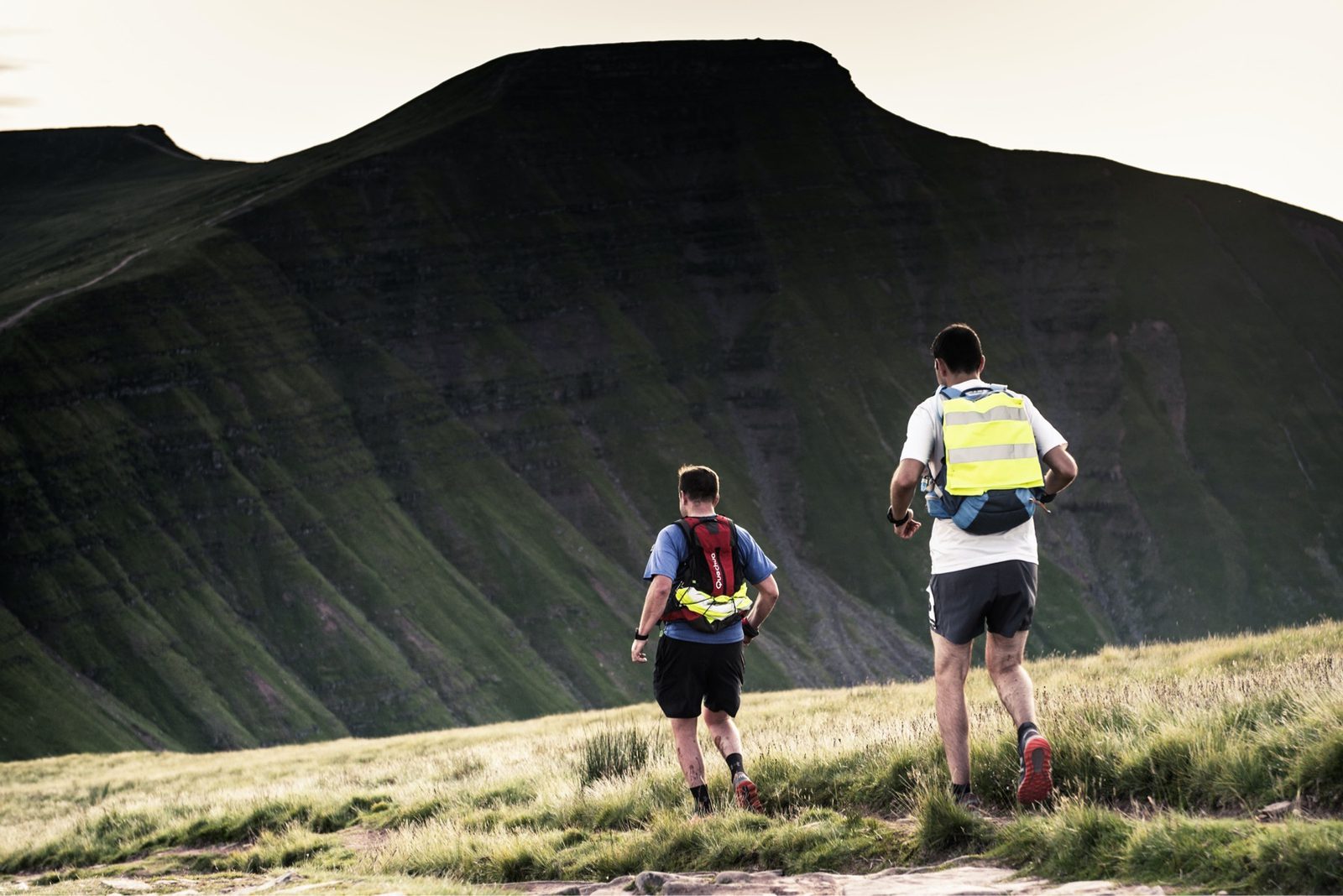 Two men trail running on a grassy mountain path, one wearing a blue shirt and red backpack, the other in a white shirt and yellow vest. A large, steep mountain with a flat top looms in the background, under an overcast sky.