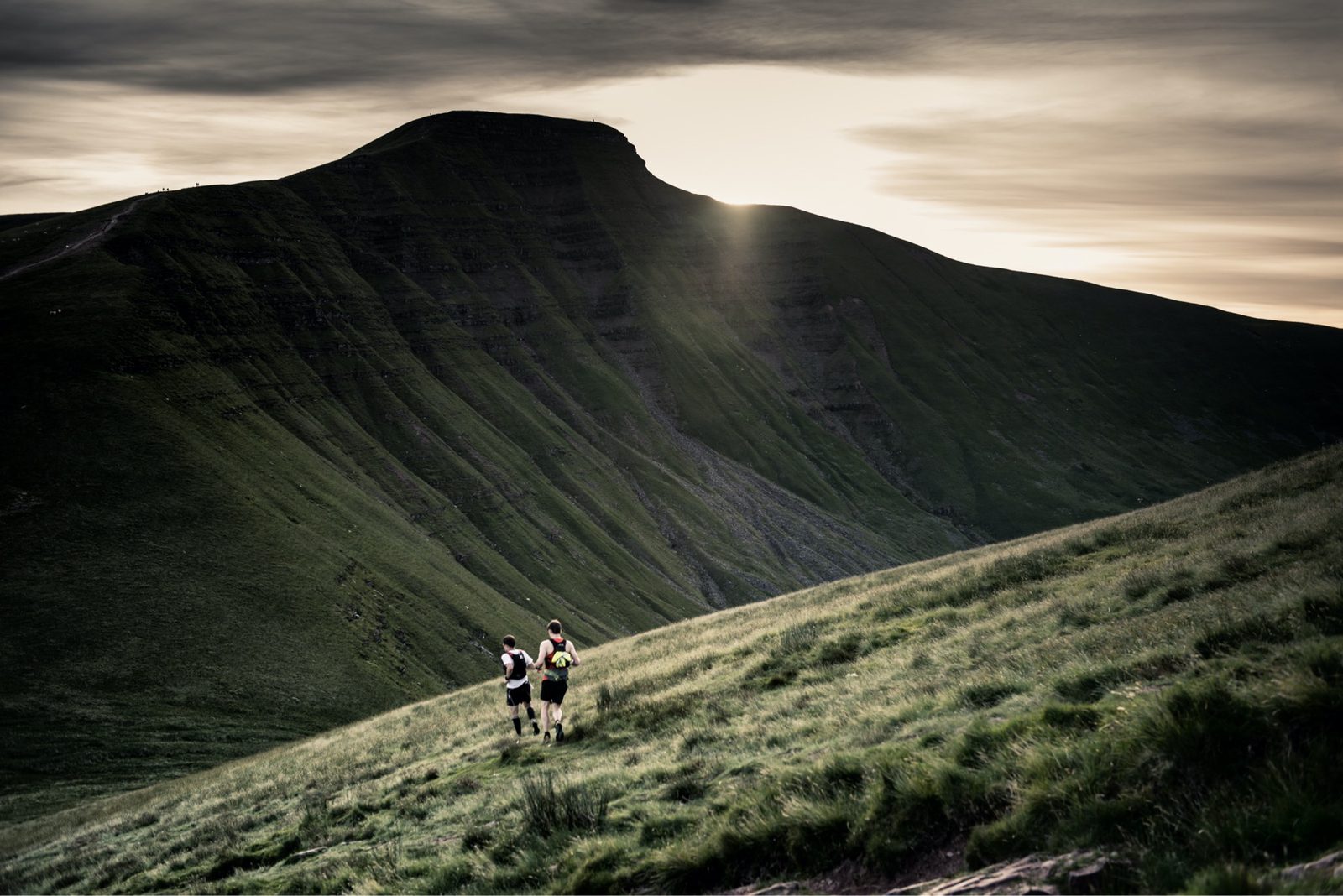 Two people run up a grassy hillside with a large, shadowed mountain in the background, partly illuminated by the setting sun. The sky is overcast, creating a dramatic, serene atmosphere.