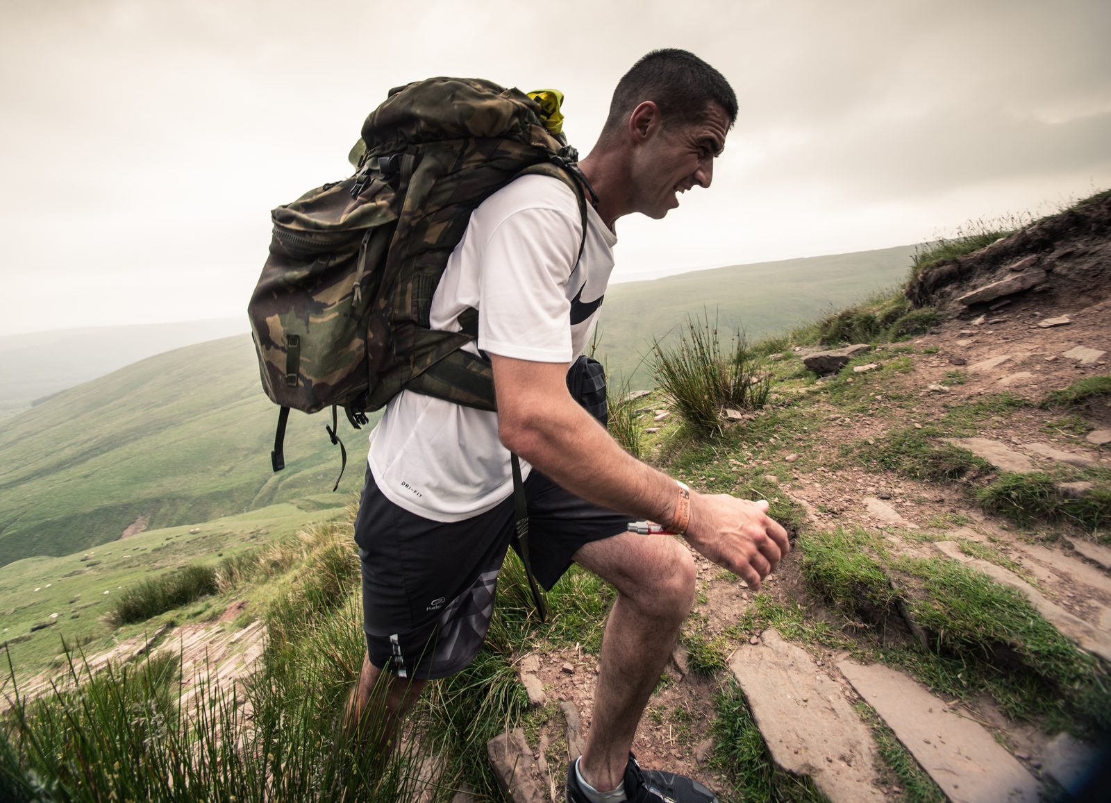 A man wearing a white t-shirt and black shorts is hiking uphill on a rocky path with a large camouflage backpack. The grassy landscape stretches out behind him under a cloudy sky. He looks determined and focused as he ascends.