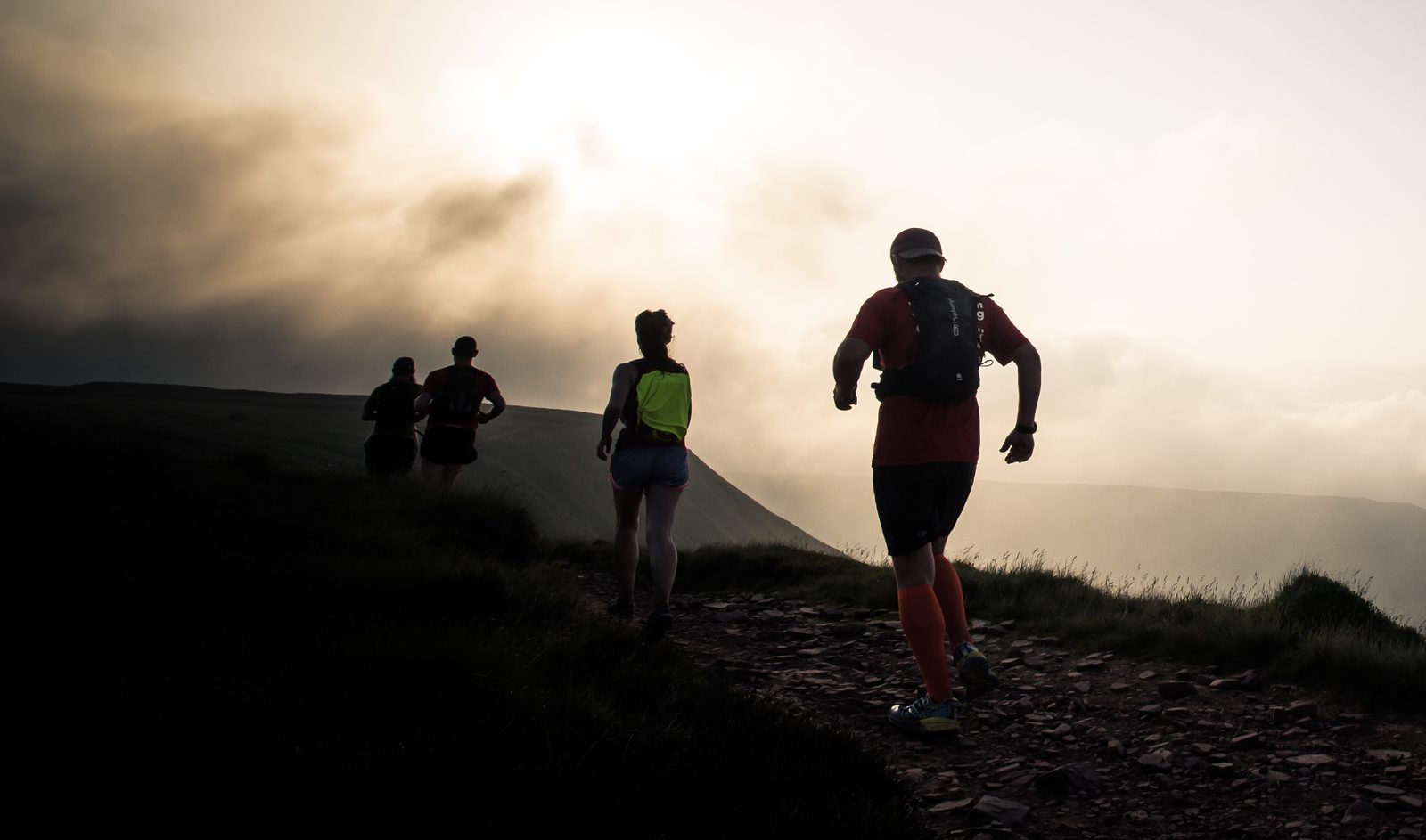 Four runners in athletic outfits, including bright tops and arm warmers, are jogging on a rocky path during sunset. The sky is filled with clouds, creating a dramatic and serene backdrop as they head towards the horizon.