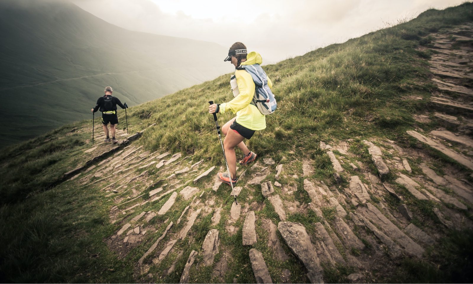 Two hikers traverse a rocky mountain trail. The foreground shows a woman in a yellow jacket and helmet, descending carefully. Another hiker, donning a black outfit, uses trekking poles to navigate uphill. Misty mountains loom in the background under a cloudy sky.