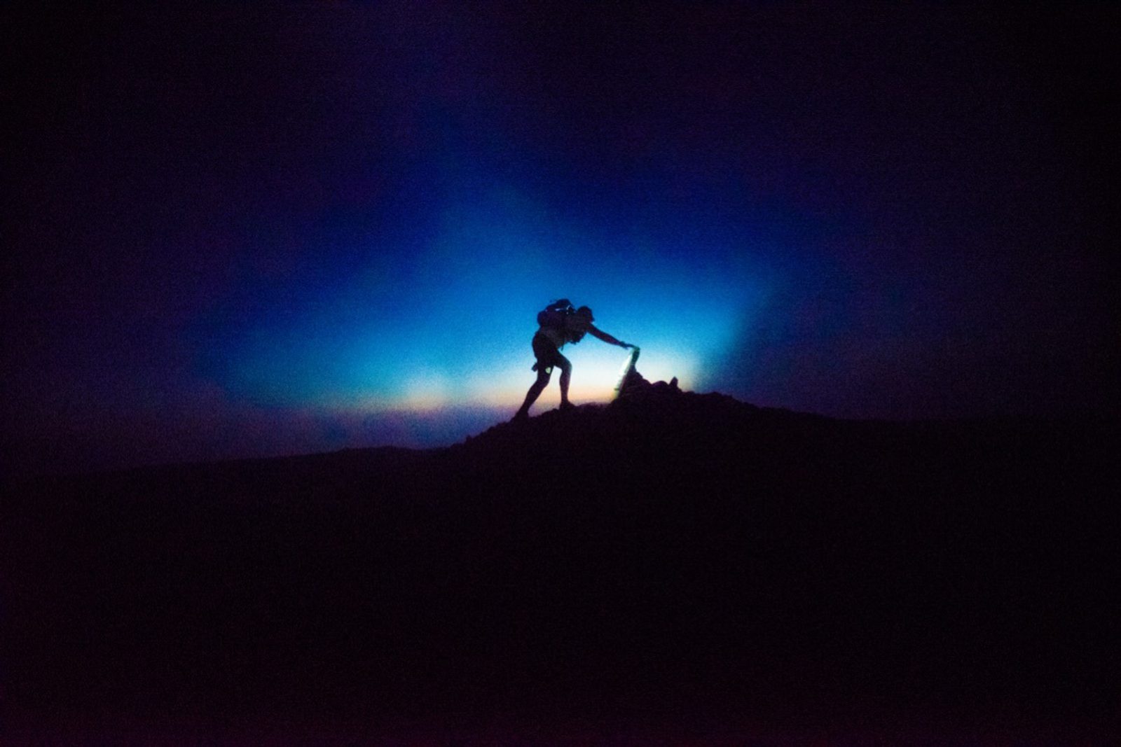 Silhouette of a person hiking up a hill at night, with a bright, blue-tinged light casting a glow behind them, creating a striking contrast against the dark sky.