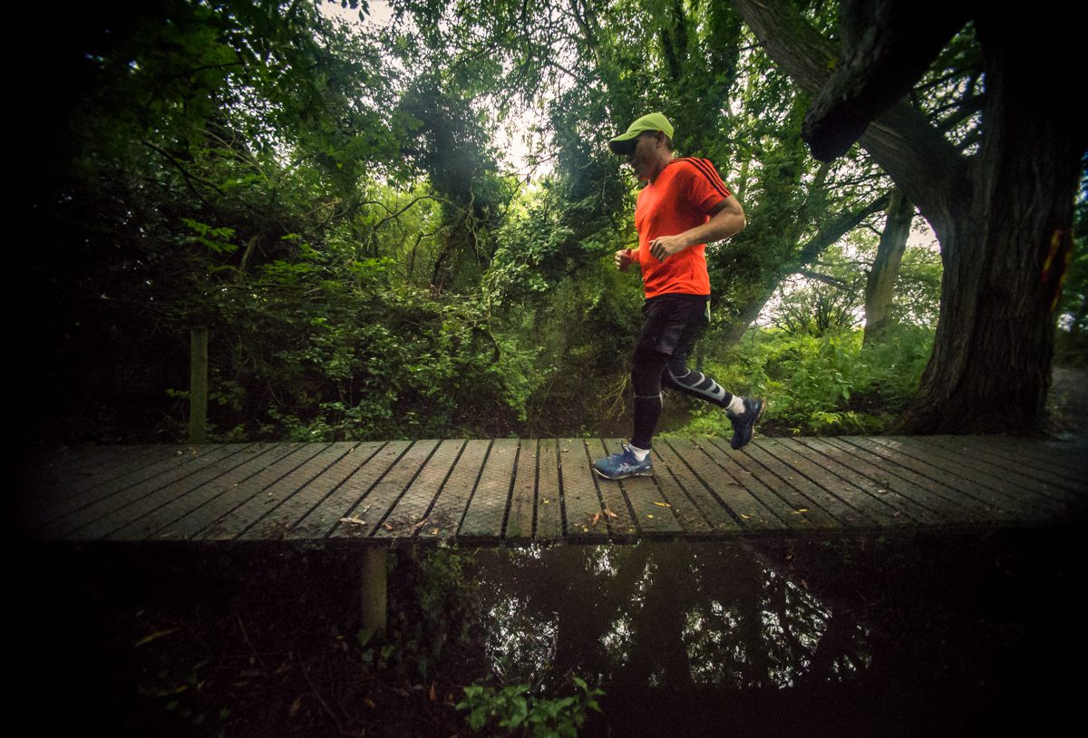 A person jogs across a narrow wooden bridge in a lush, green forest. They are wearing a bright orange shirt, black leggings with a pattern, blue running shoes, and a green cap. The scene is reflected in the calm water below the bridge.