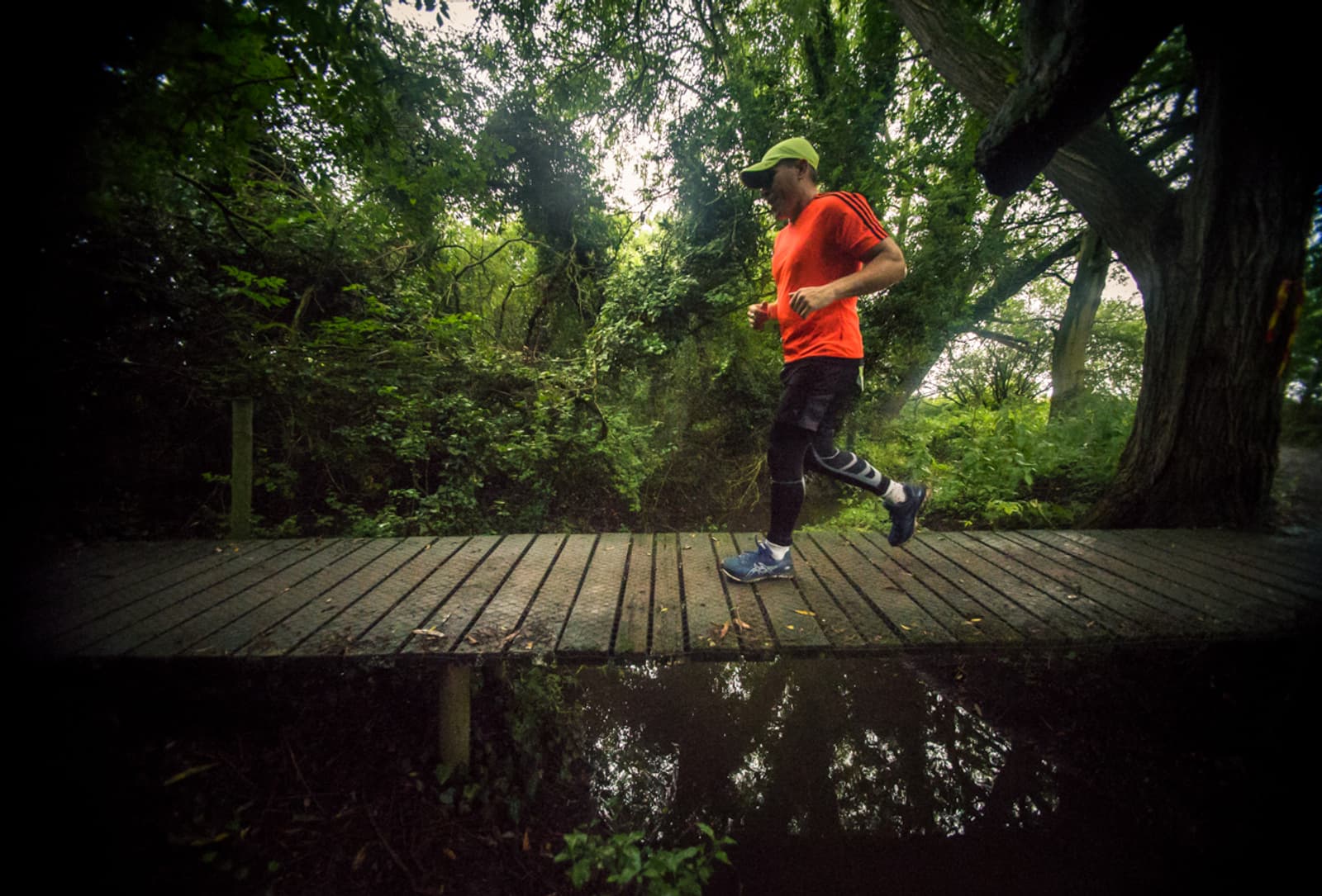 A person jogs across a narrow wooden bridge in a lush, green forest. They are wearing a bright orange shirt, black leggings with a pattern, blue running shoes, and a green cap. The scene is reflected in the calm water below the bridge.