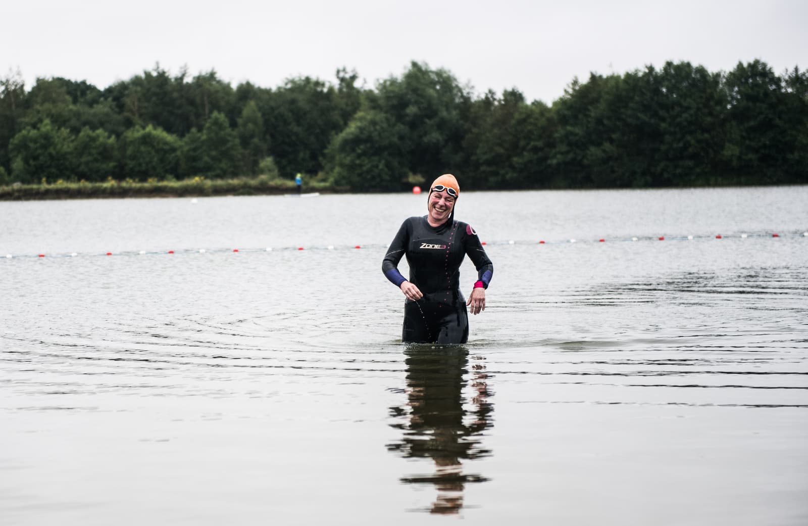 A person wearing a wetsuit and swim cap is standing in a lake with water up to their thighs, smiling. Trees and buoy markers are visible in the background under an overcast sky.