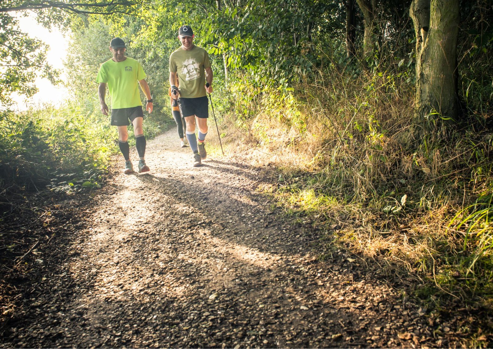 Two men are walking on a sunlit dirt path surrounded by greenery. The man on the left wears a bright yellow t-shirt and black shorts, while the man on the right wears a beige t-shirt and dark shorts and walks with a crutch. Sunlight filters through the trees.